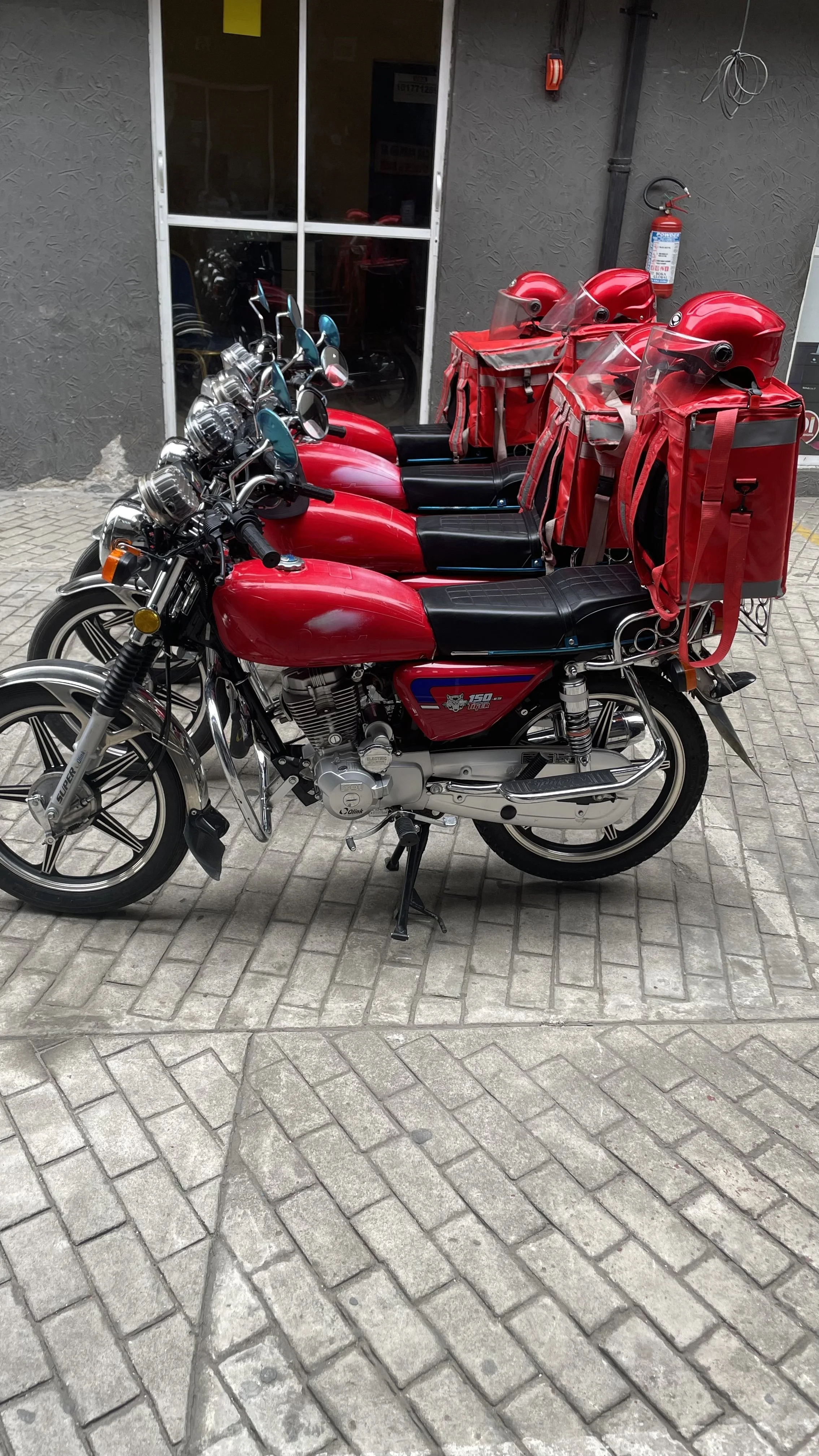Line of three red motorcycles with delivery backpacks parked outside a building with a dark gray wall and a glass door.
