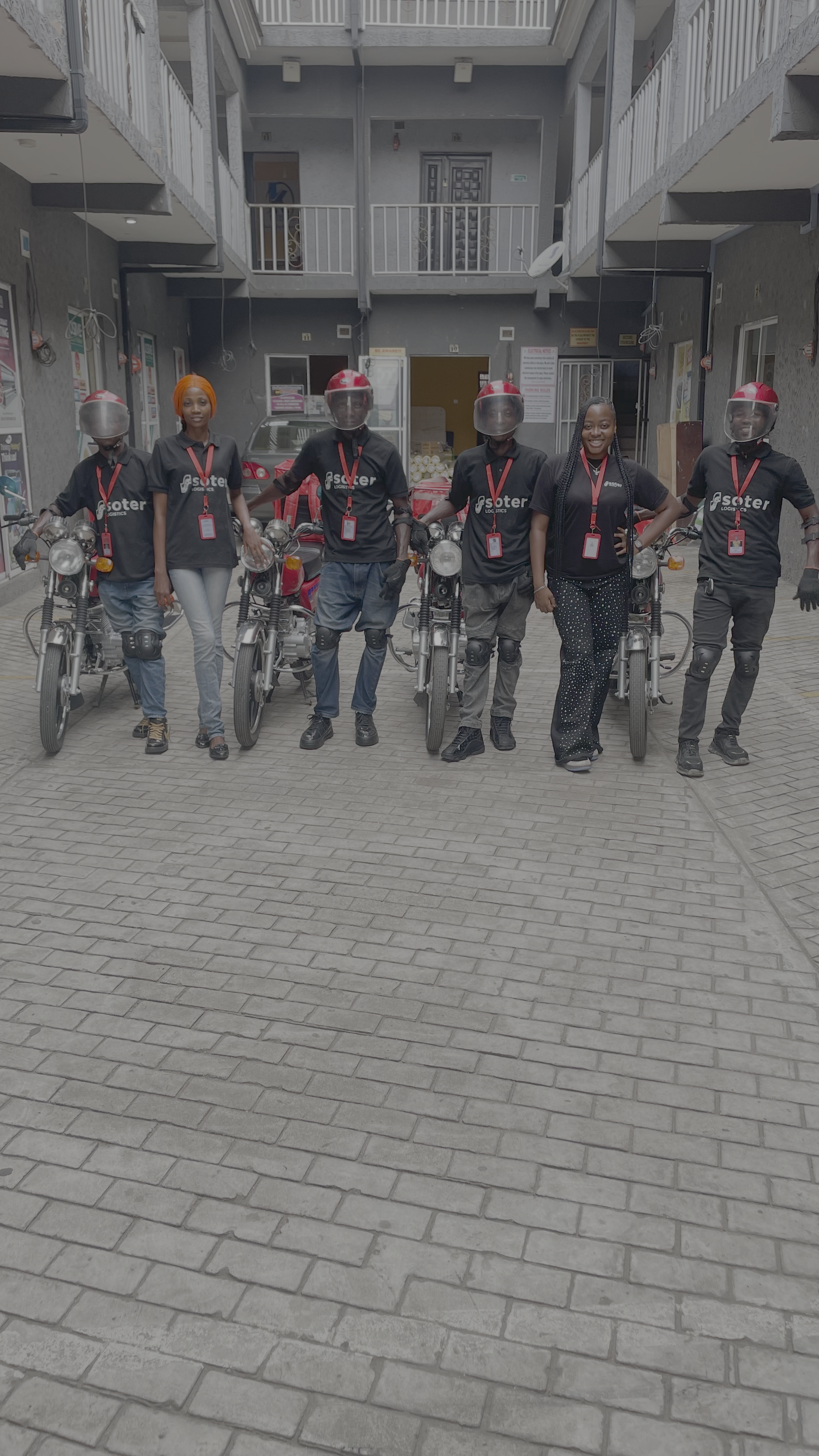 Group of six people standing in a courtyard with three motorcycles, one woman, and five men wearing black t-shirts with 'Scooter Logistics' printed on them, some wearing helmets, some with gloves, and one woman dressed in black with white polka dots.