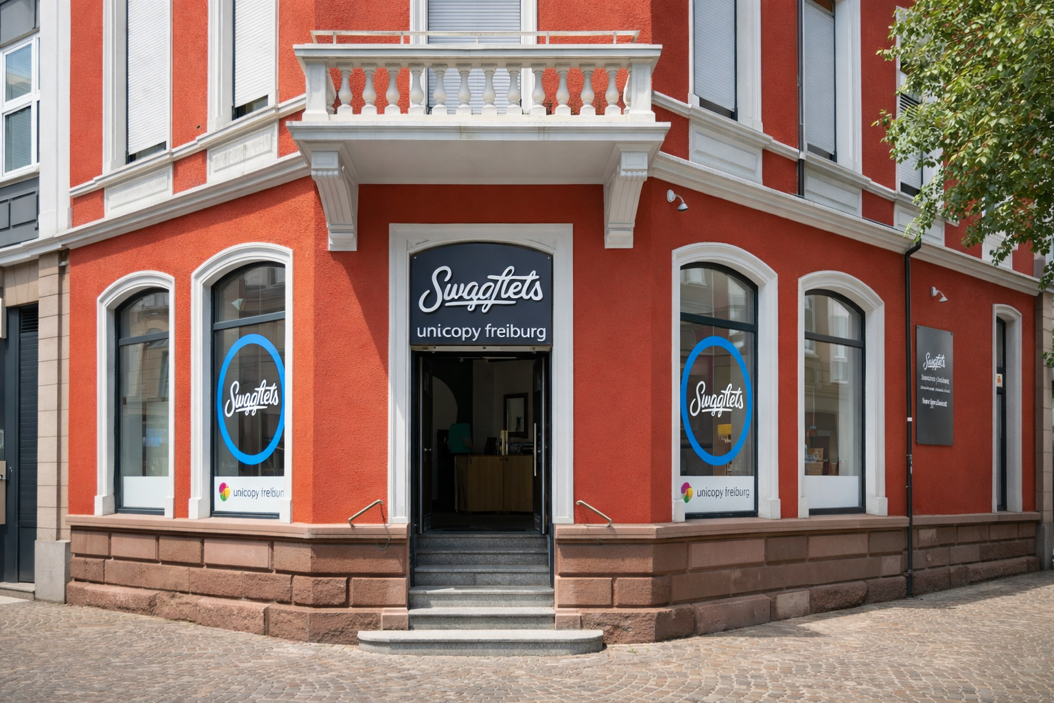 Ein rotes Gebäude mit weißen Fenstern und Türen, das einen Laden namens 'Swaglets' zeigt. Der Laden befindet sich in Freiburg und hat große Glasschaufenster mit blauer Umrandung. Es gibt eine Treppe, die zum Eingang führt.