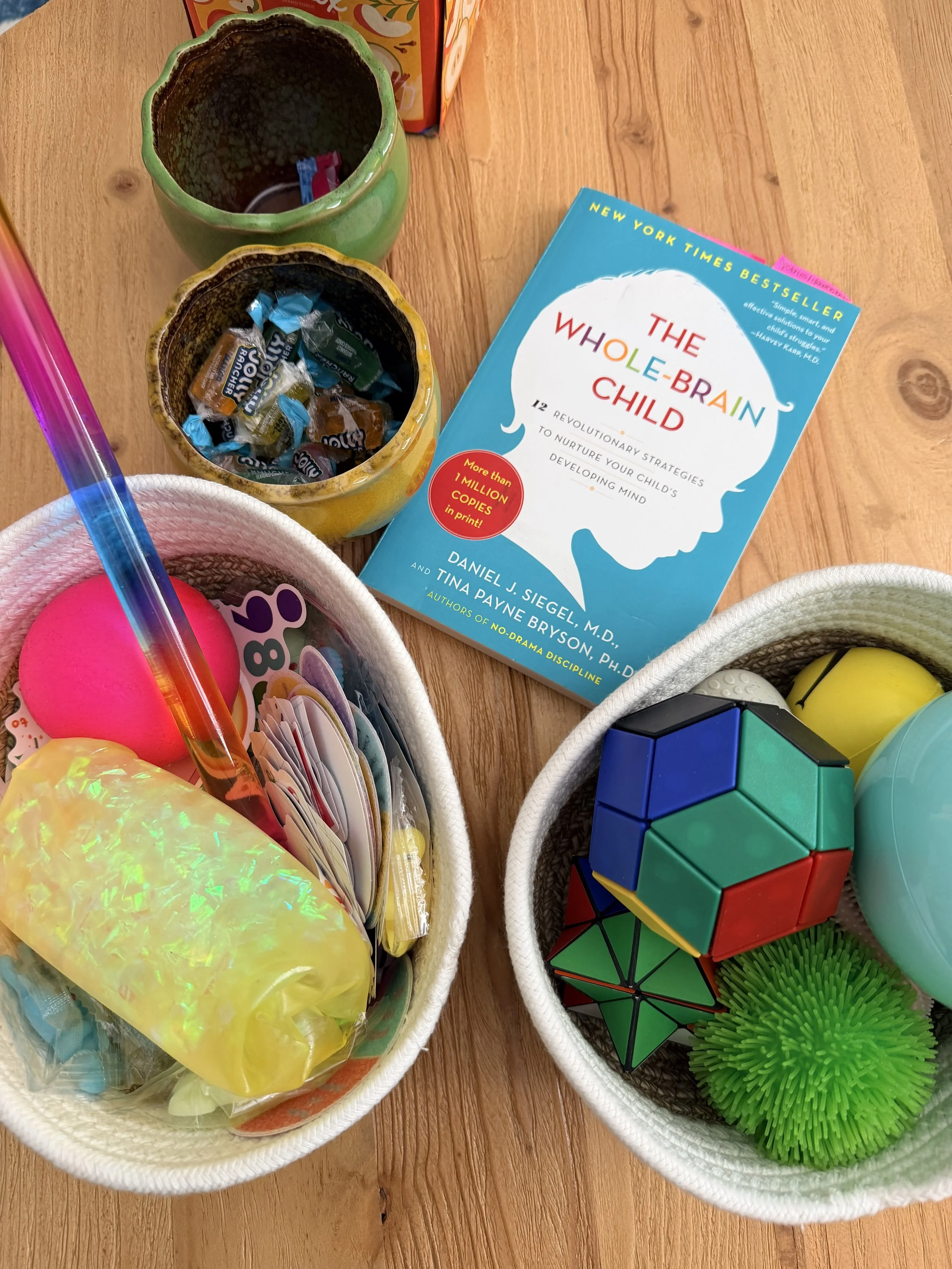 A wooden table displaying a children's book titled 'The Whole-Brain Child', two baskets filled with toys and candies, and two decorated cups.