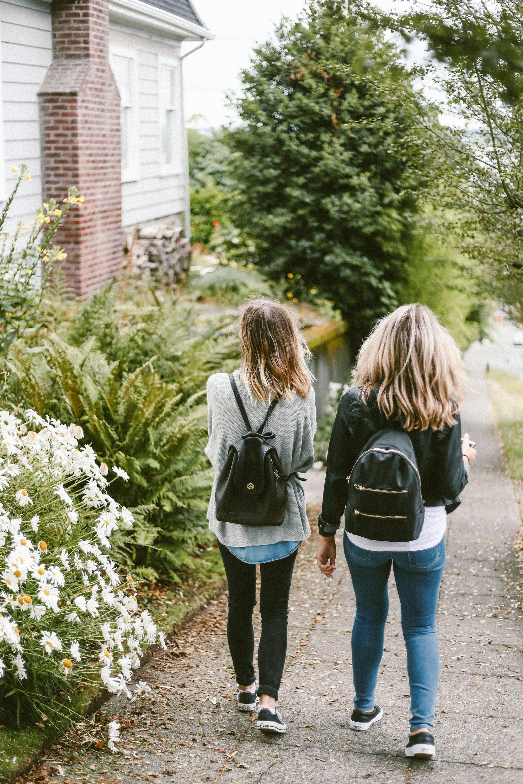 Two young women walking along a sidewalk next to a garden with white daisies and green foliage, surrounded by trees and a house with white siding and a brick chimney.