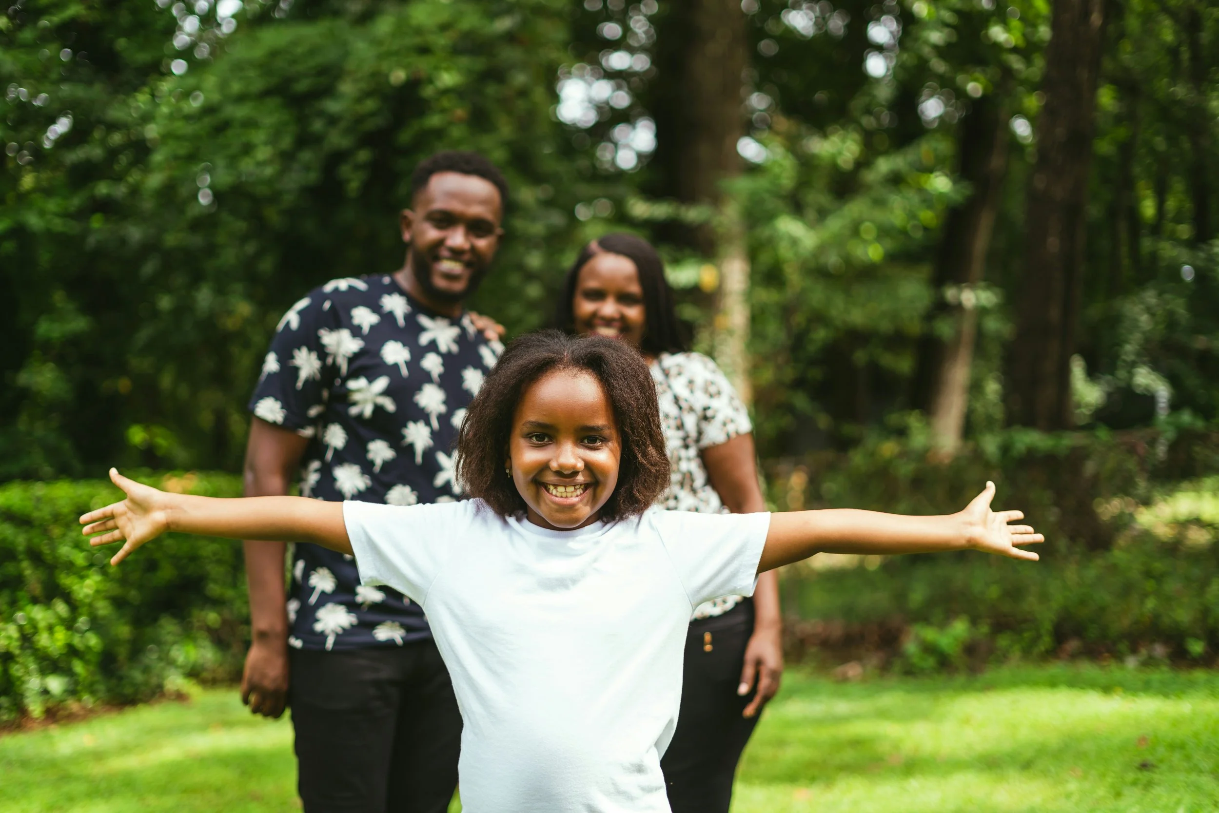 A young girl with outstretched arms smiling at the camera, with an African American family of three in the background outdoors in a lush, green park.