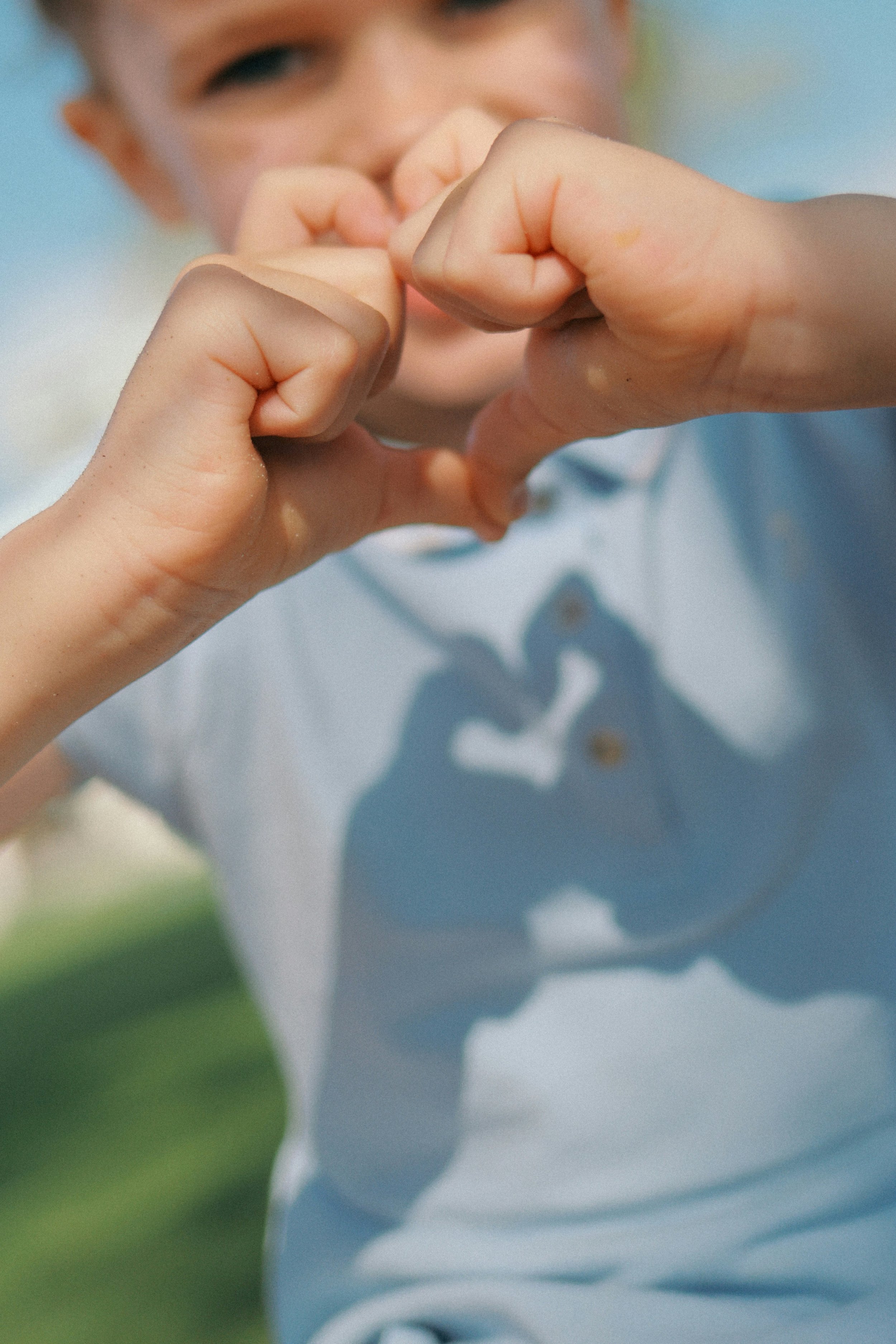 Person forming a heart shape with hands, smiling behind. Blurred greenery and blue sky in the background.