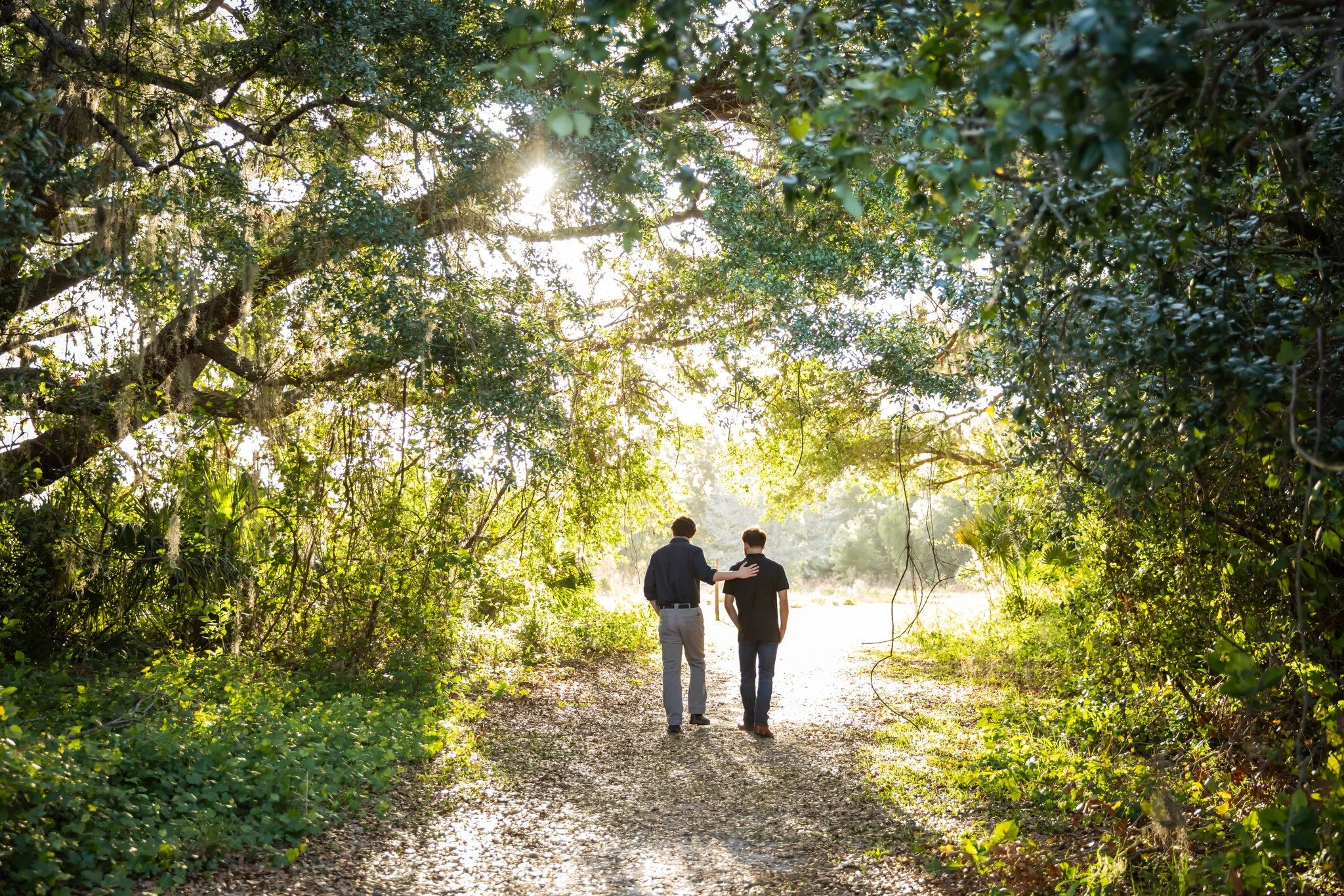 Two people, one in a black shirt and jeans, the other in a dark blazer and light pants, walking along a shaded trail in a lush, green forest with sunlight filtering through the trees.