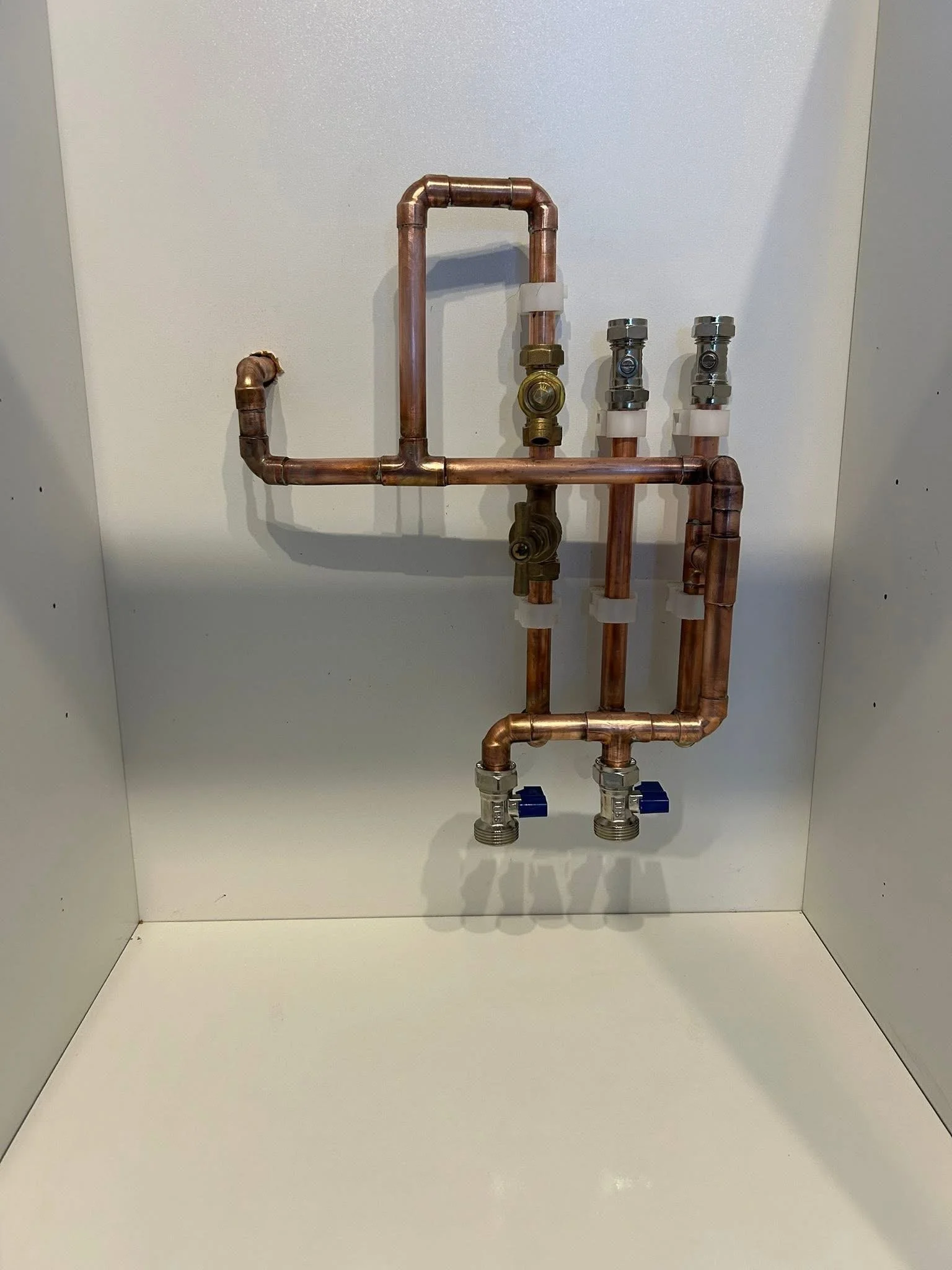 Arrangement of copper plumbing pipes with shut-off valves and fittings mounted on a white enclosed wall.