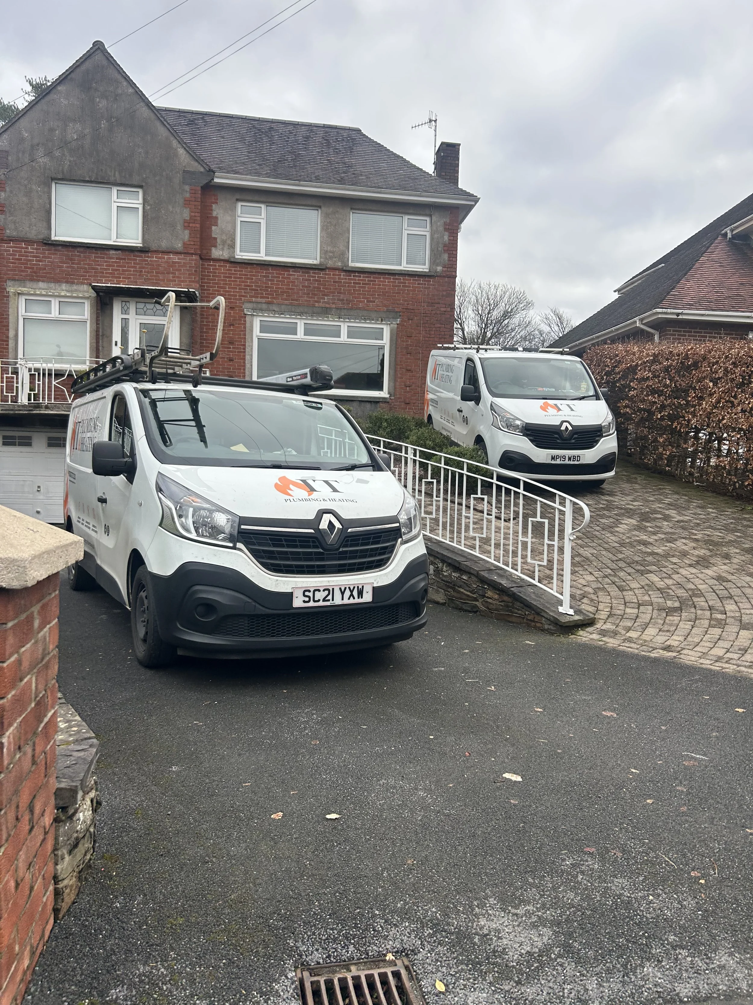 Two plumbing and heating service vans parked outside a house with bushes and a driveway.