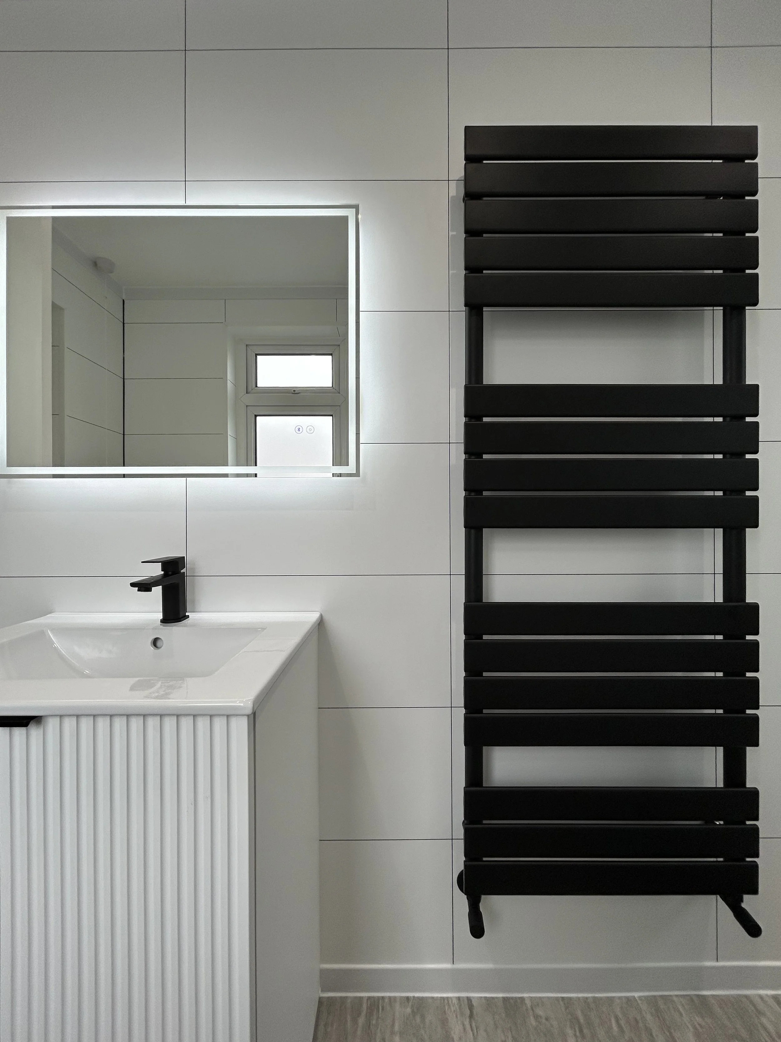 A modern bathroom scene with a white sink and black faucet, a rectangular mirror with LED lighting, a small window, and a black heated towel radiator mounted on a white tiled wall.