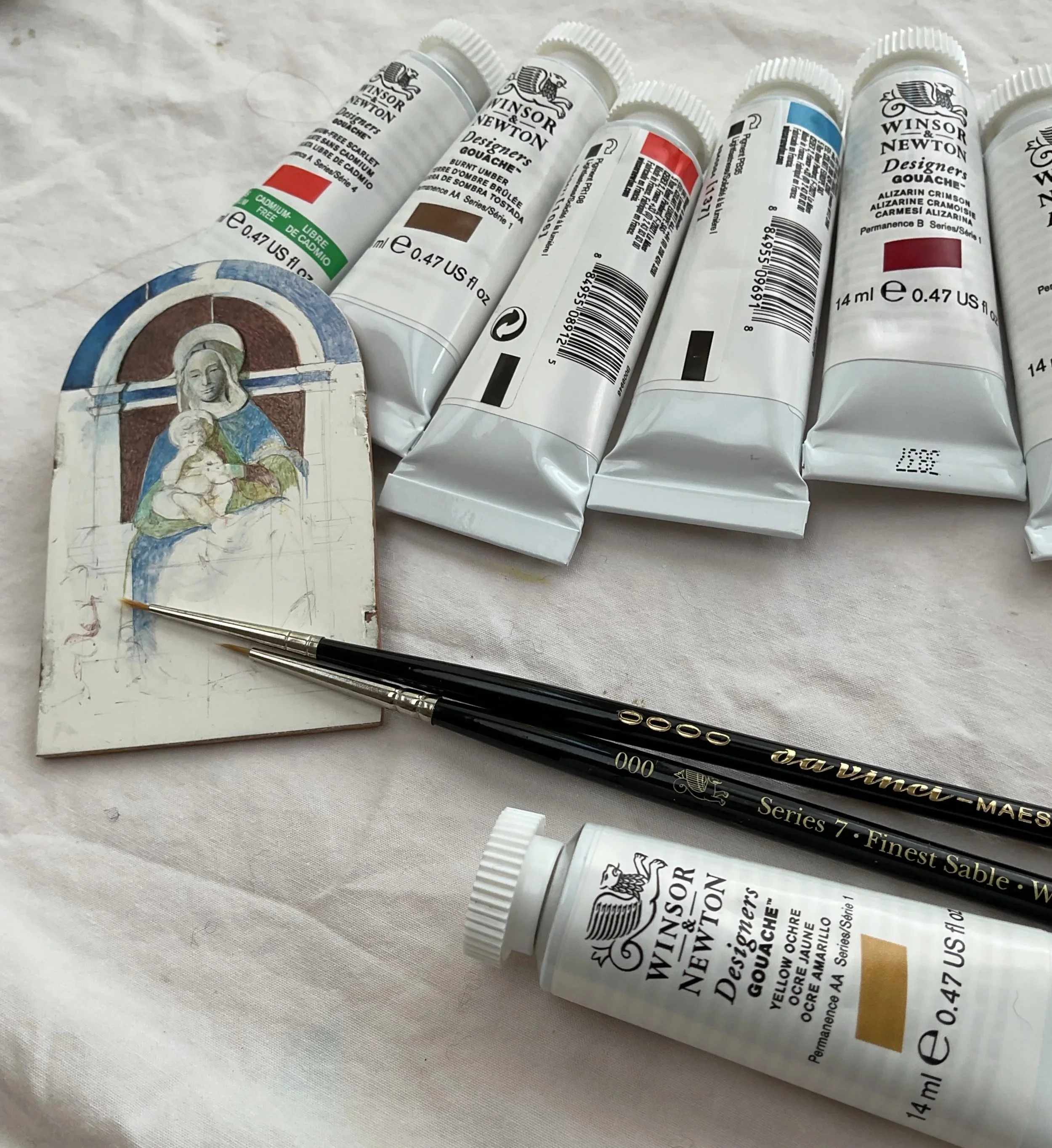 Several tubes of Windsor & Newton Designer's Gouache paint lying on a white surface, with a partially painted religious icon image on a small board nearby.