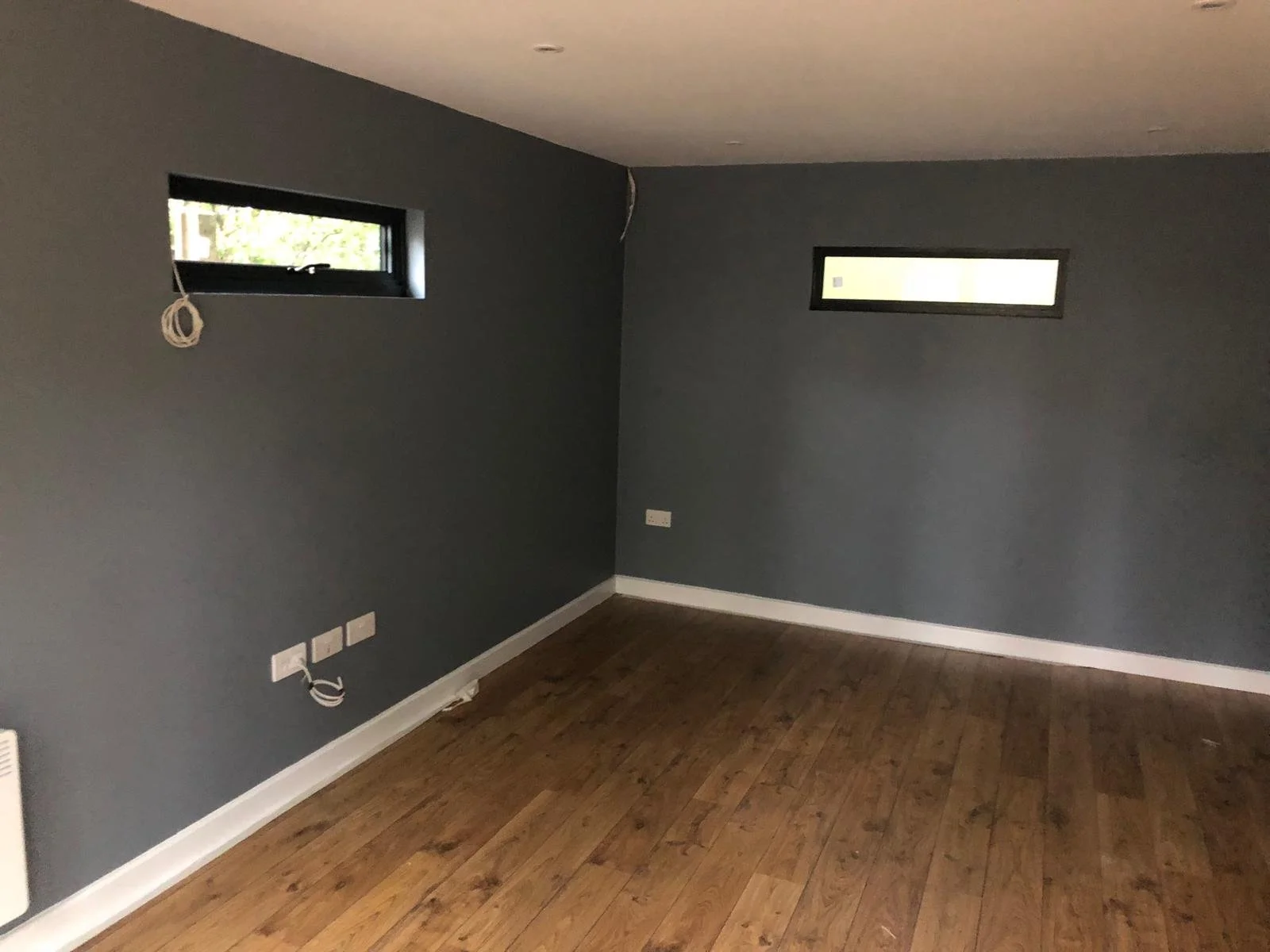 Empty room with dark gray walls, hardwood floor, two small horizontal windows, and electrical outlets with cables.