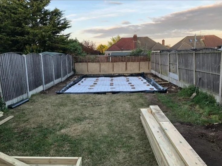 A backyard under construction, with a grid of tiles and a black liner at the far end, surrounded by wooden and metal fences, with some wooden planks in the foreground and houses with rooftops in the background.