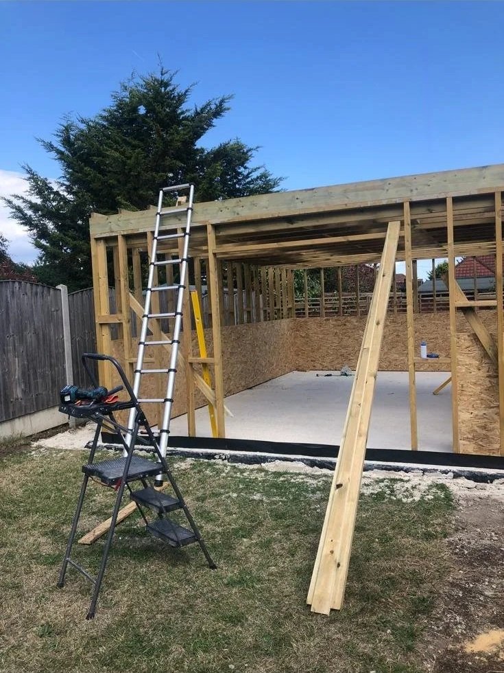 Construction of a wooden shed or garage with a concrete foundation, surrounded by a backyard with a wood fence and trees. There is a ladder, a step stool, and construction tools in the scene.