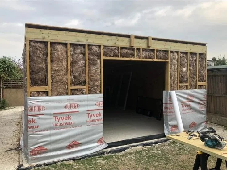 A small building under construction with frame and insulation visible, partially covered with Tyvek house wrap. Construction tools are on a table nearby.