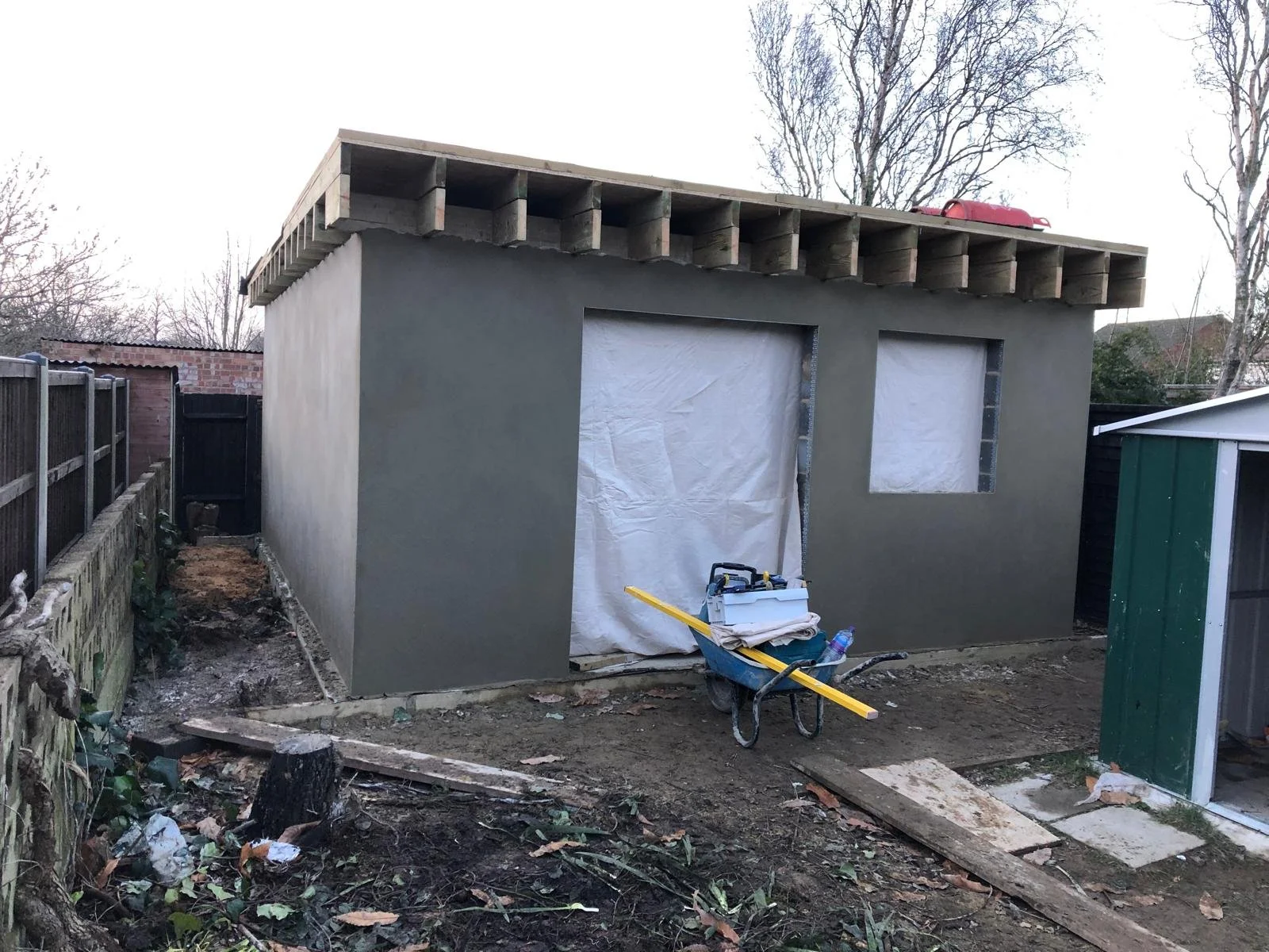 A small building under construction with a partially finished exterior wall, two window openings covered with white material, and construction tools in front. The roof frame is exposed.