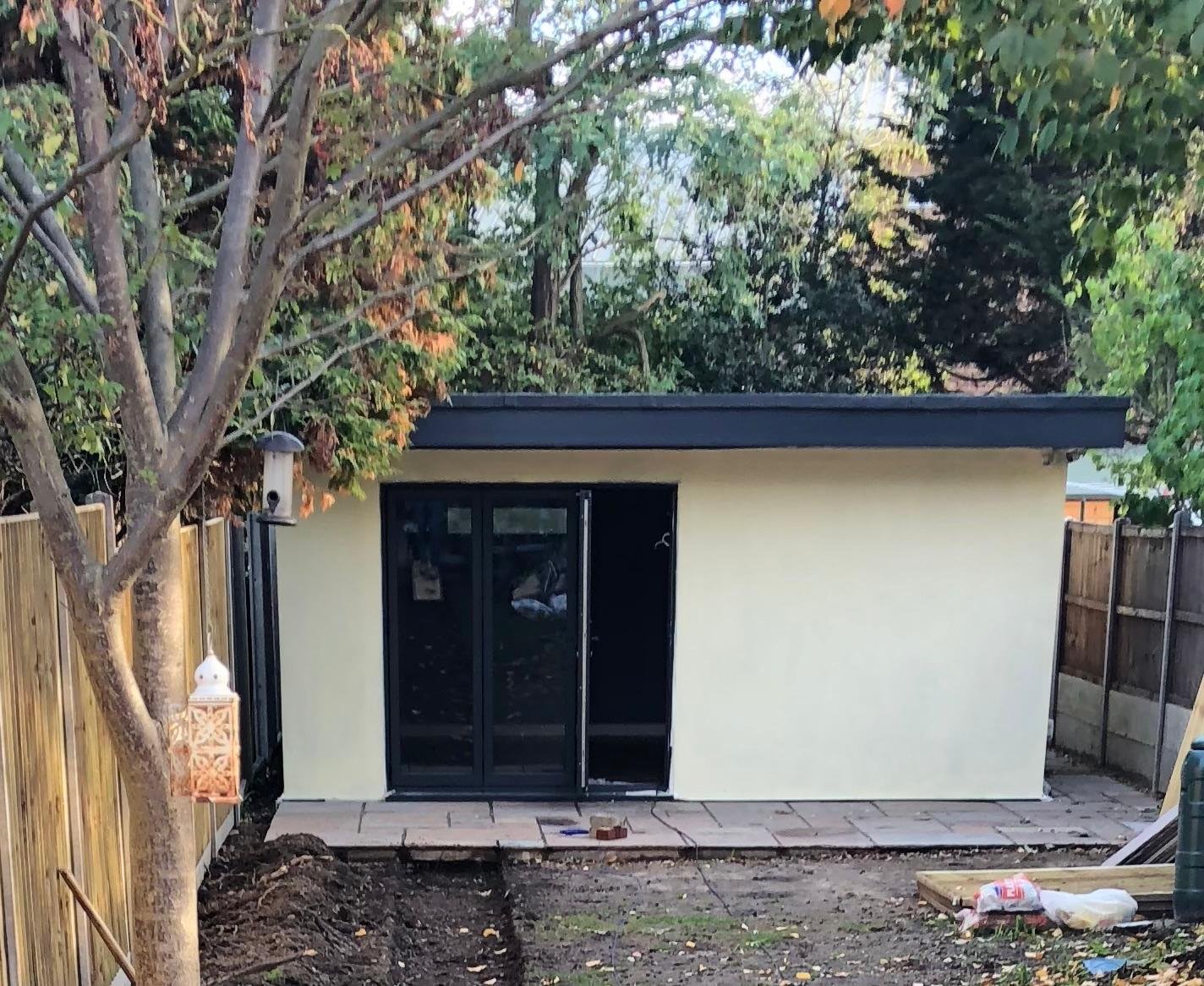 A small backyard structure with a flat black roof, beige walls, and black sliding glass doors, surrounded by a wooden fence and trees with green and orange leaves, and some construction materials on the ground.