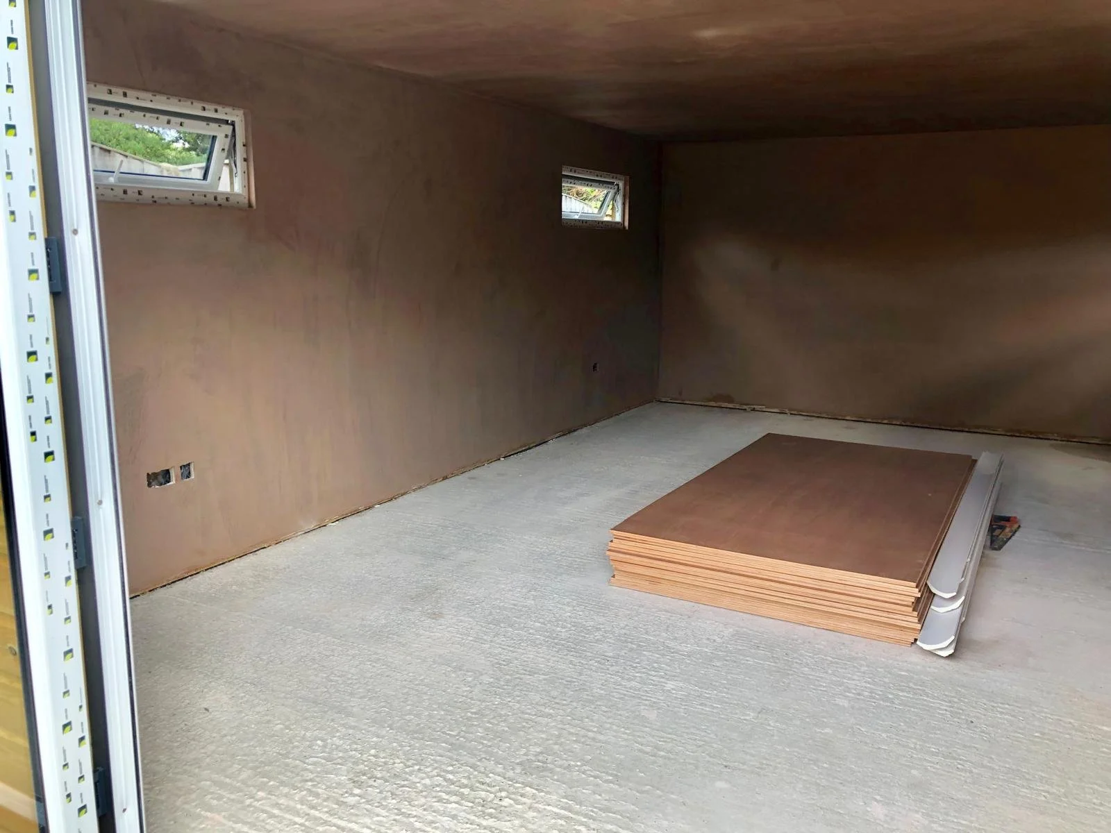 Interior of a room under construction with two small windows, brown drywall on the walls, and a vacuum-sealed stack of plywood sheets on the floor.