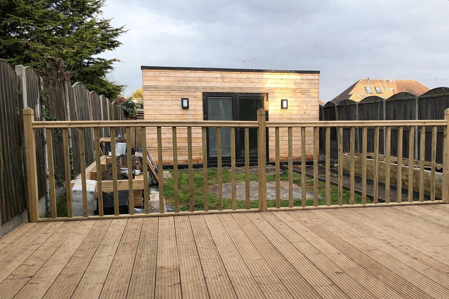 New wooden deck with safety rail overlooking a small backyard with patchy grass, a wooden shed under construction, and neighboring houses in the background. Overcast sky.