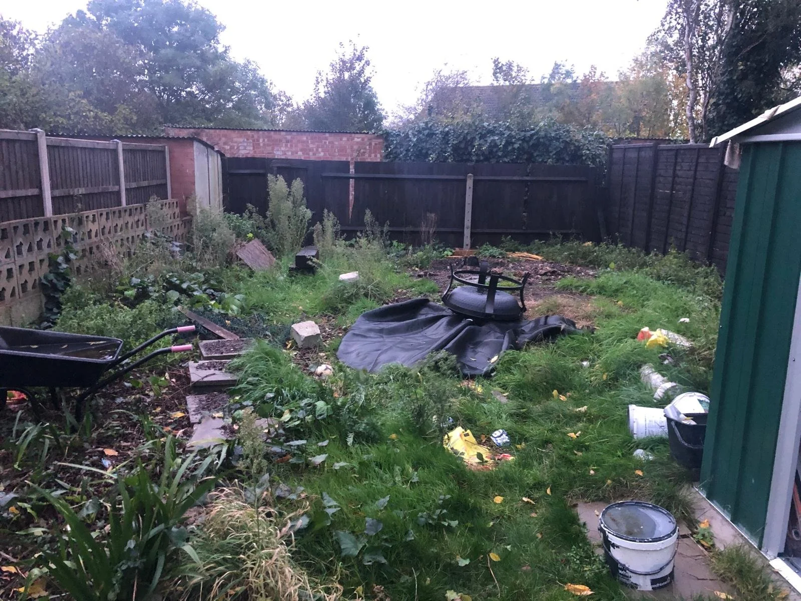 A messy backyard with overgrown grass, scattered debris, a black tarp, a wheelbarrow, and discarded containers surrounded by a wooden fence.