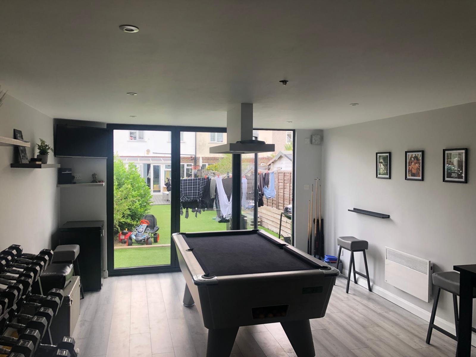 Interior of a modern game room with a pool table, dumbbells, and a view of a backyard with outdoor laundry drying and a lawn.