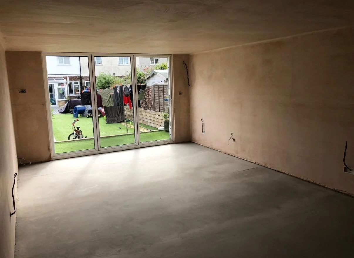 Empty room with unfinished walls and floor, large glass sliding door leading to a backyard with lawn, clothes hanging on a line, and outdoor objects like a bicycle and a washing machine.