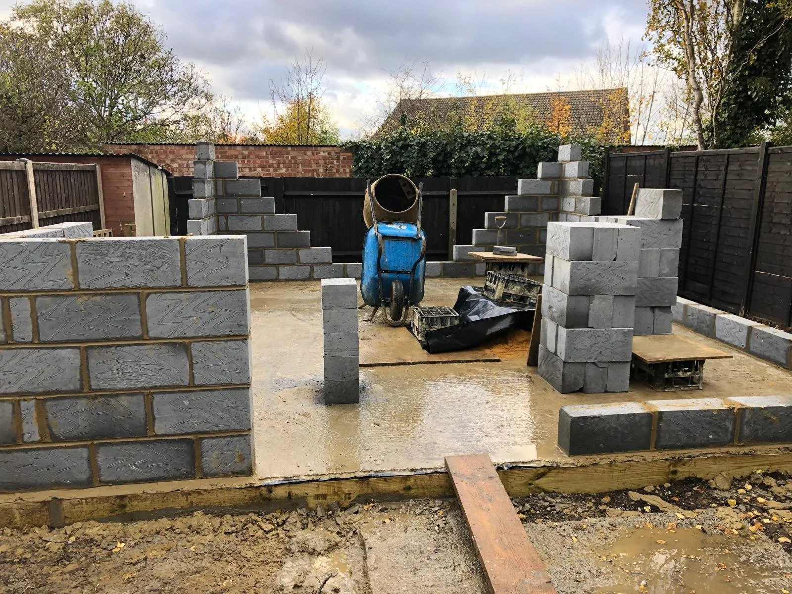 Construction site with partially built brick walls, a wheelbarrow, and construction tools, on a concrete foundation with muddy ground.