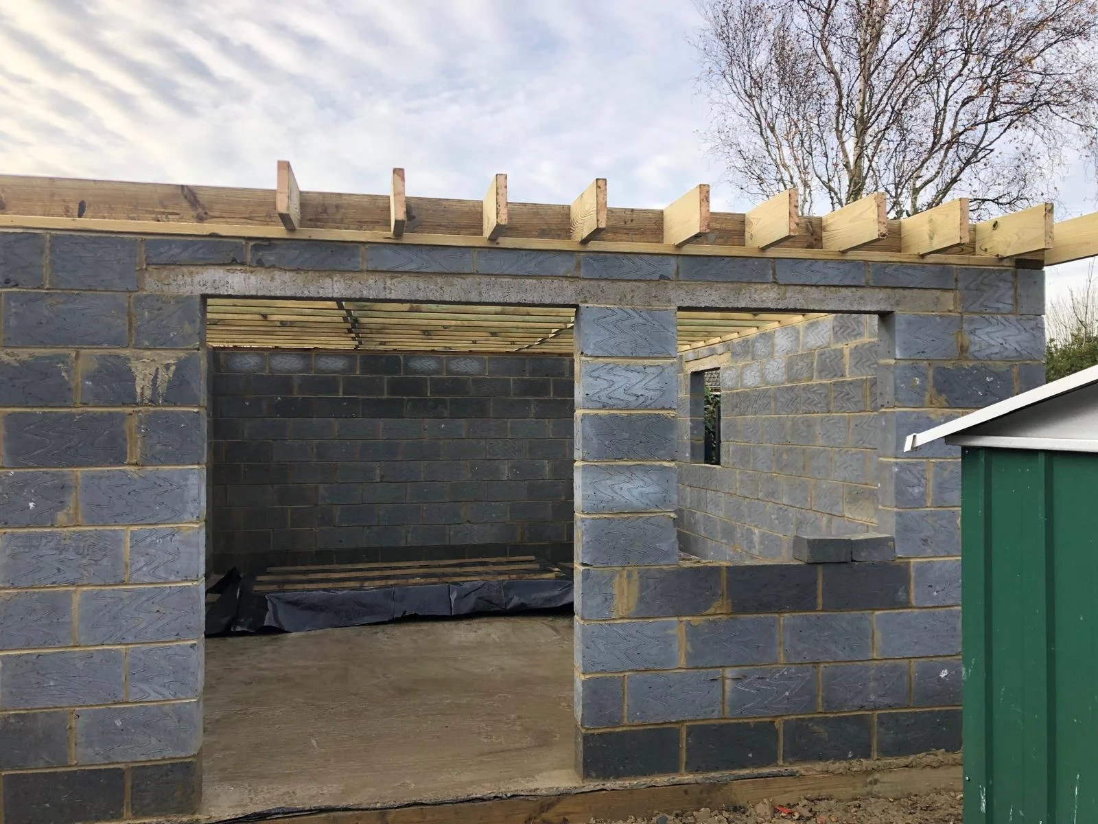 The image shows a construction site with a partially built brick structure. The structure has an opening for a door and a window, with wooden beams on top for the roof framework. The sky is partly cloudy with some trees visible in the background, and