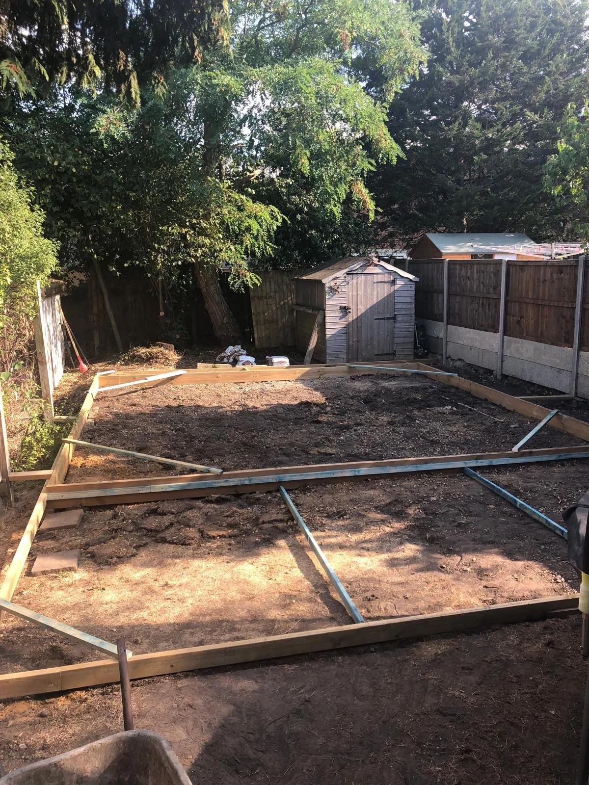A backyard garden area under construction with a rectangular foundation frame made of wooden planks and metal supports, surrounded by dirt, with a shed and trees in the background, and a wooden fence along the perimeter.