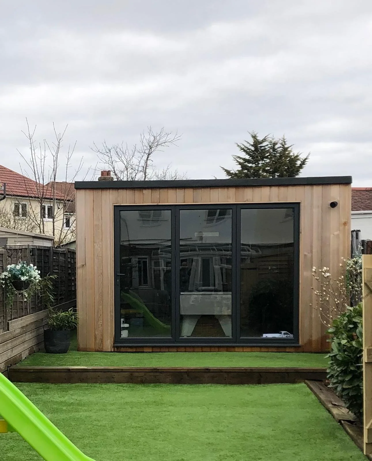 A small modern wooden garden house with large glass sliding doors, set on a raised wooden platform in a backyard with well-maintained green lawn and decorative plants.