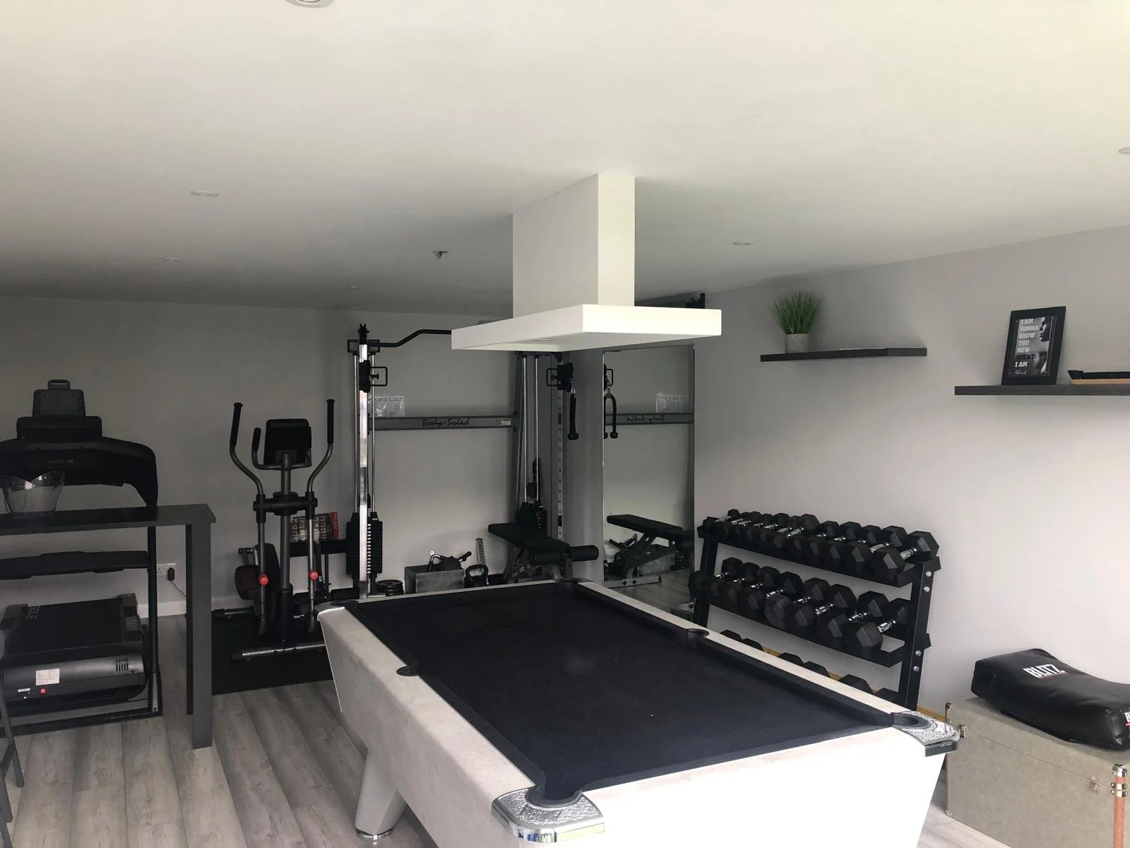 Home gym with workout equipment including a treadmill, elliptical, weight rack with dumbbells, Pilates machine, and a pool table, all in a room with gray walls and wood flooring.