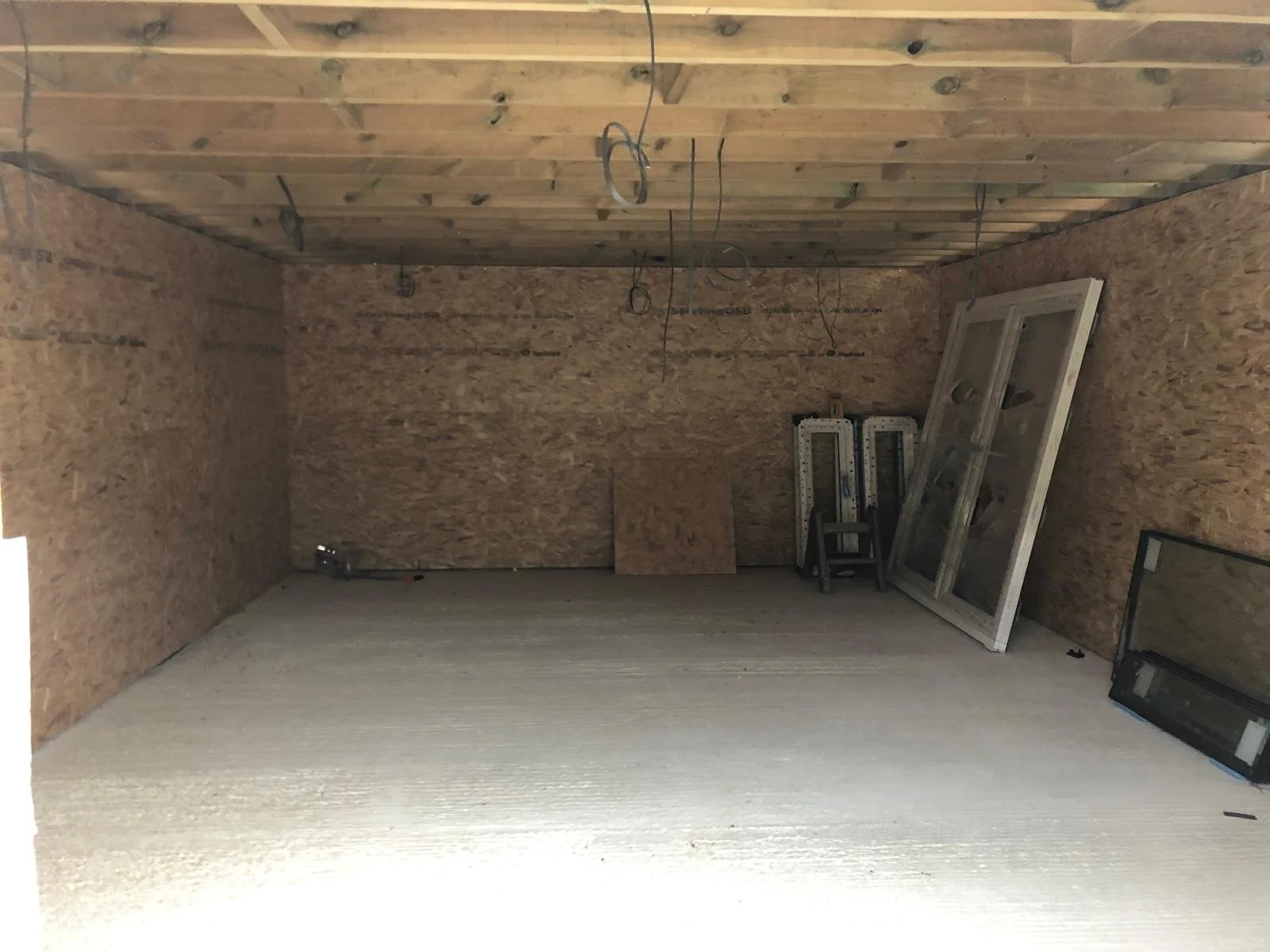 An unfinished room under construction with plywood walls and ceiling, and a concrete floor. There are wires hanging from the ceiling, a window frame leaning against the wall, and building materials or tools like ladders and panels.