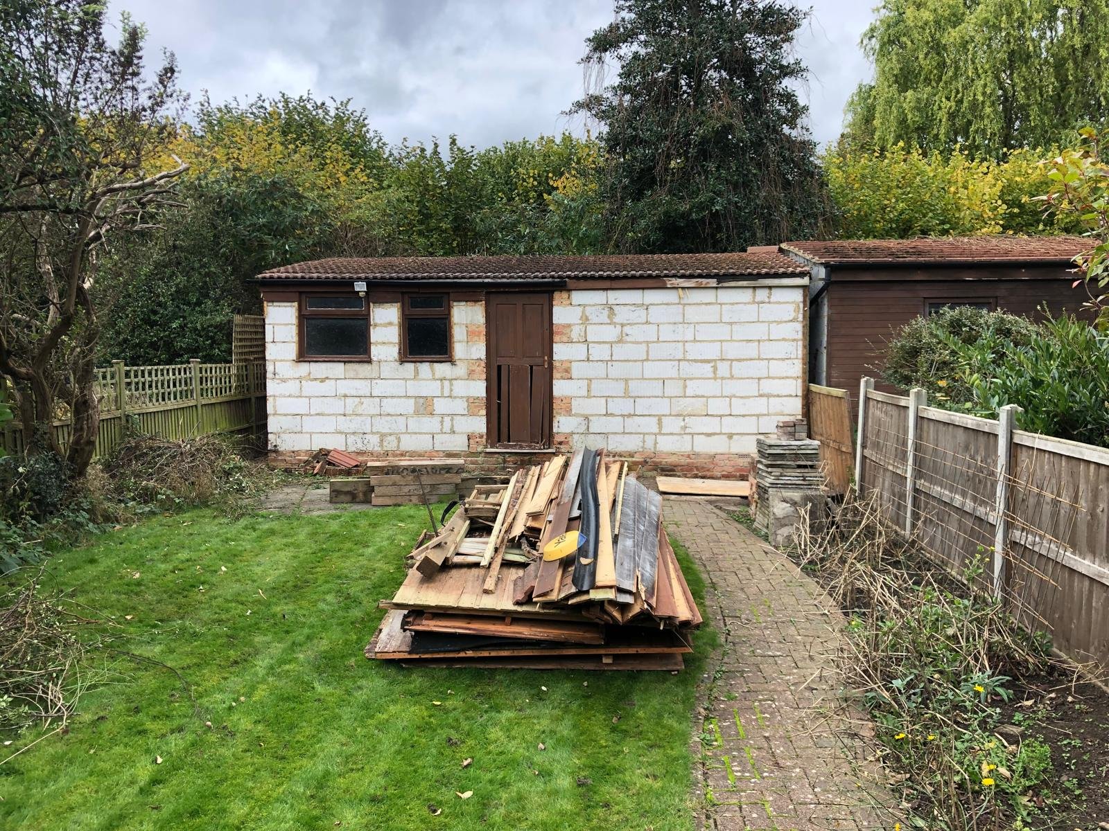 Backyard scene showing a small, unfinished building with a wooden door and two windows, surrounded by a wooden and wire fence, with piles of wooden and metal materials in the foreground, grass, trees, and cloudy sky.