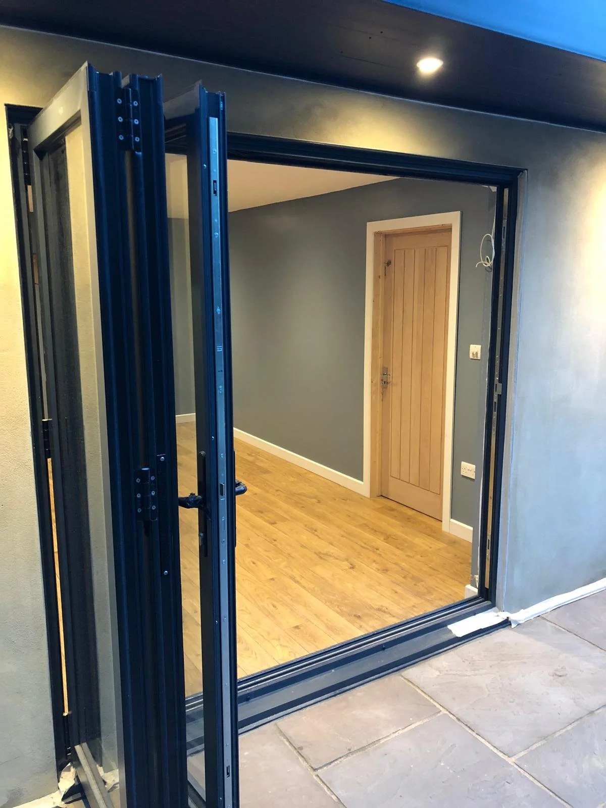 View of an interior room with a wooden door, hardwood flooring, gray walls, and a partially installed black sliding or folding door frame, seen from the outside.