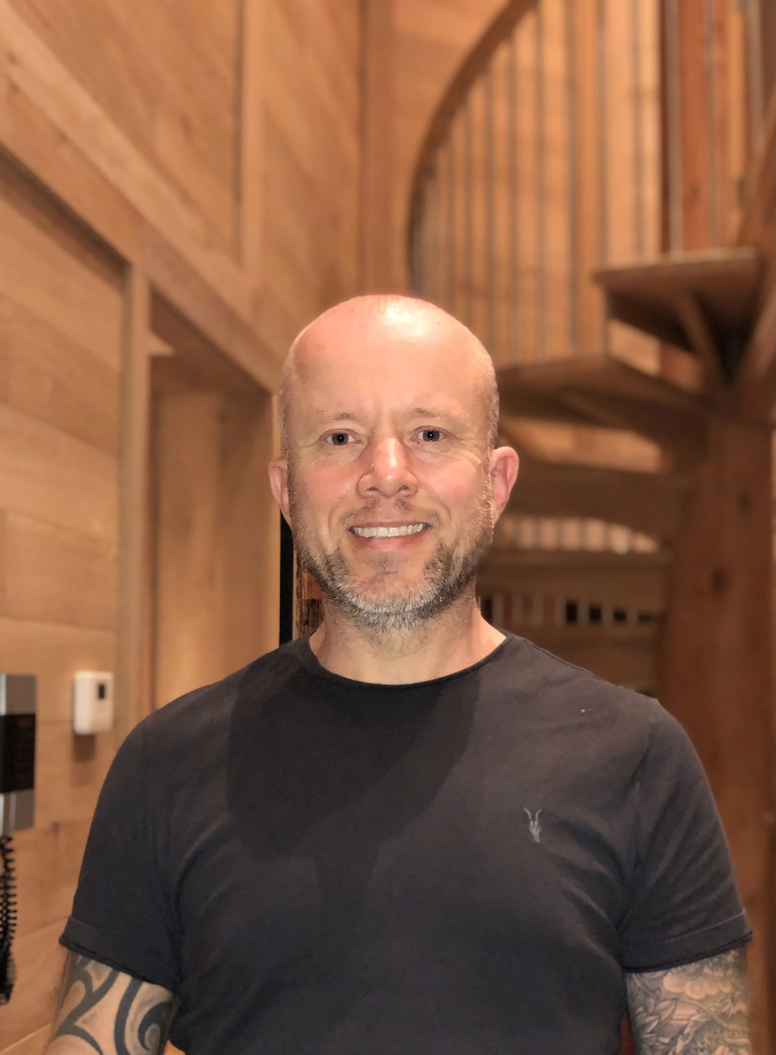 A man with a bald head and a beard, smiling, wearing a black t-shirt with tattoos on his arms, standing in a room with wooden walls and shelves.