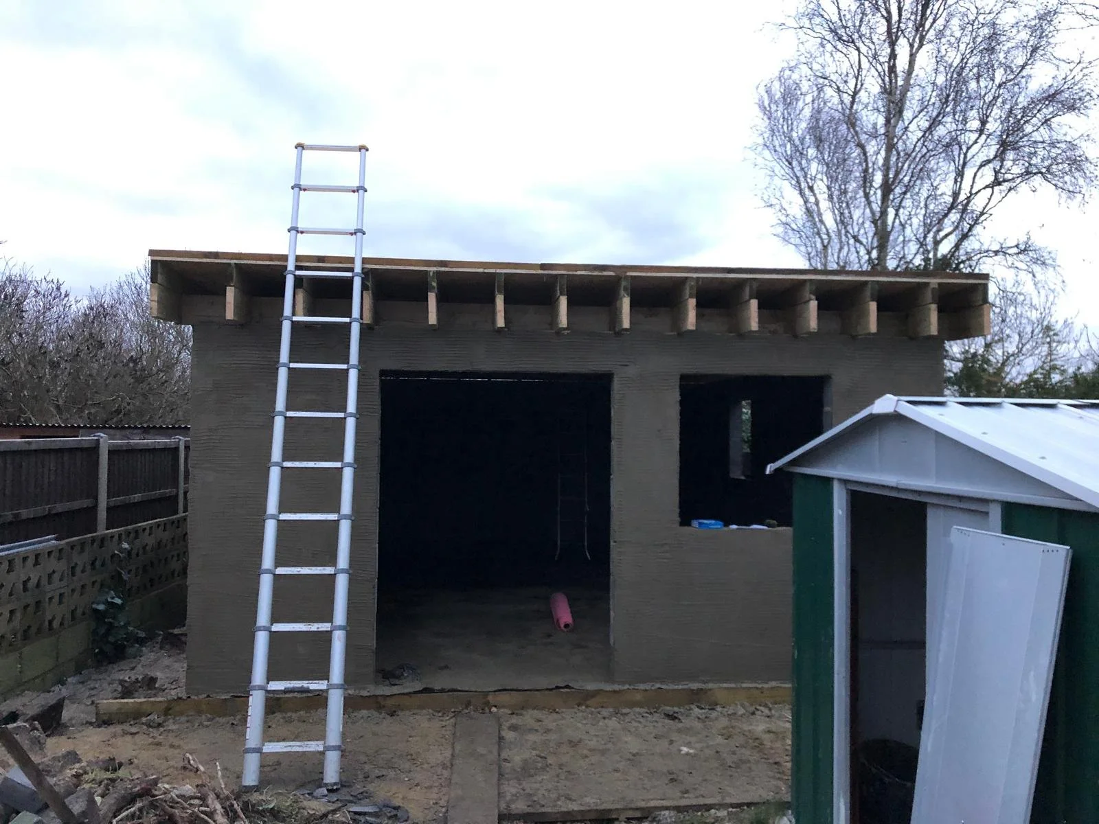A small building under construction with an open front, a ladder leaning against it, and a pink pipe inside. The upper part of the structure has exposed beams, and there is a small shed on the right side.
