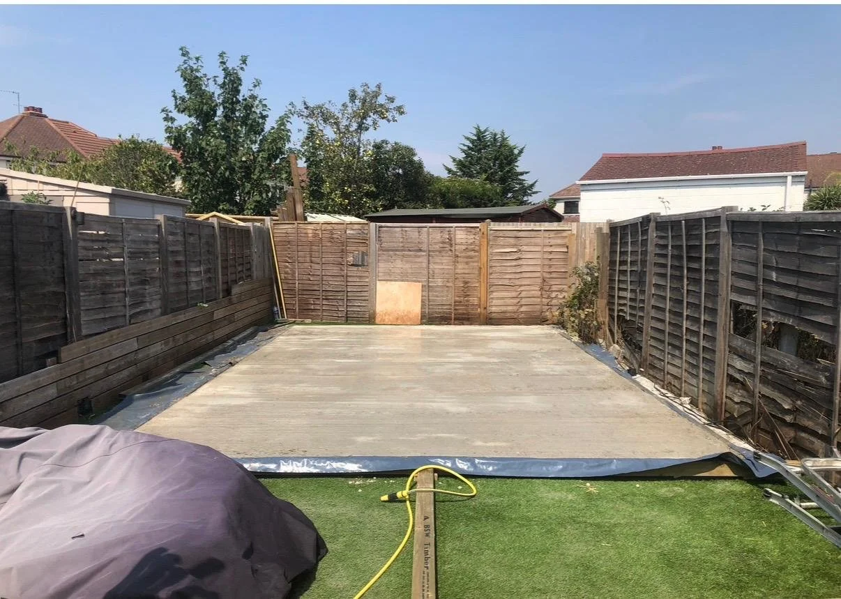 A backyard with a newly poured concrete patio, surrounded by a wooden fence. Construction tools and materials are present, and the sky is clear with some trees and neighboring houses visible in the background.