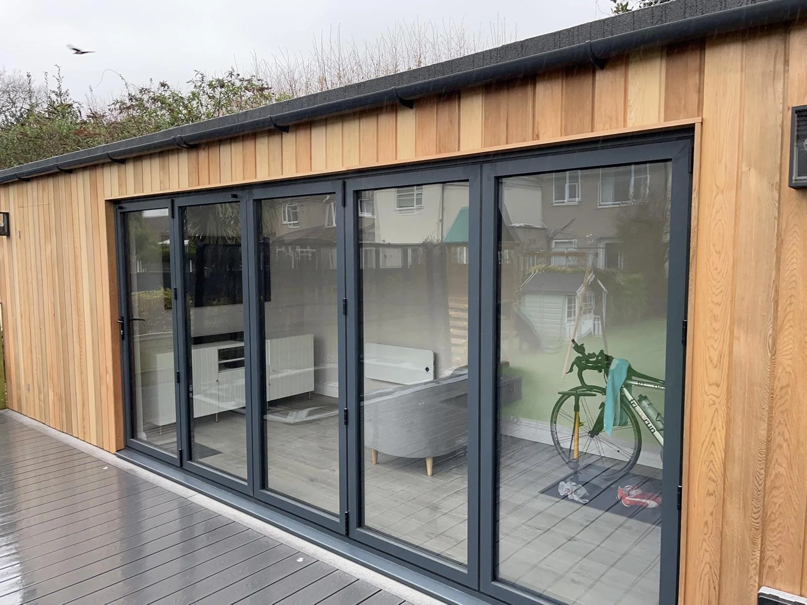 A modern wooden house with large glass sliding doors, a bicycle inside, and a deck in the foreground.