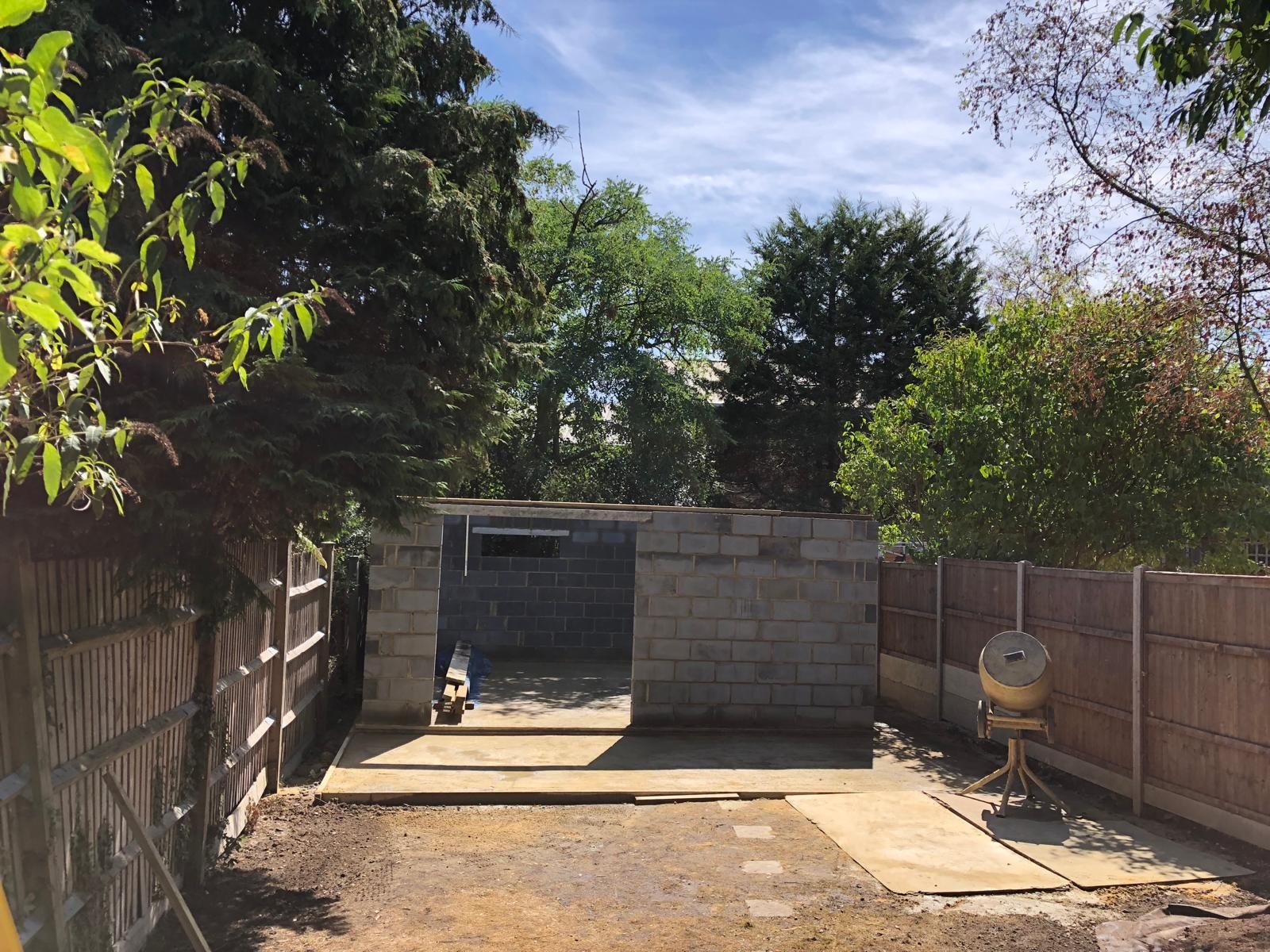 A backyard with trees, a partially built brick structure, a wooden fence, a cement mixer, and a concrete slab on the ground.