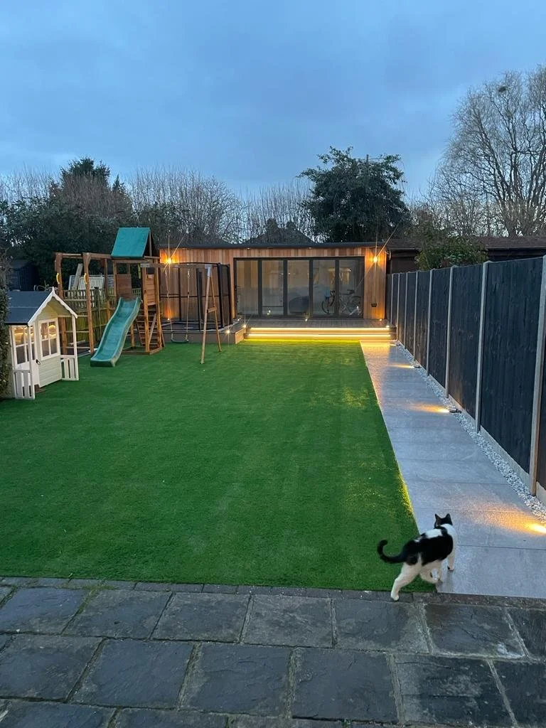 A backyard with a grassy play area, a small plastic playhouse, a wooden playset with a slide, a trampoline with a safety enclosure, and an illuminated steps leading to a glass-enclosed patio. A black and white cat walks on the paved walkway along the