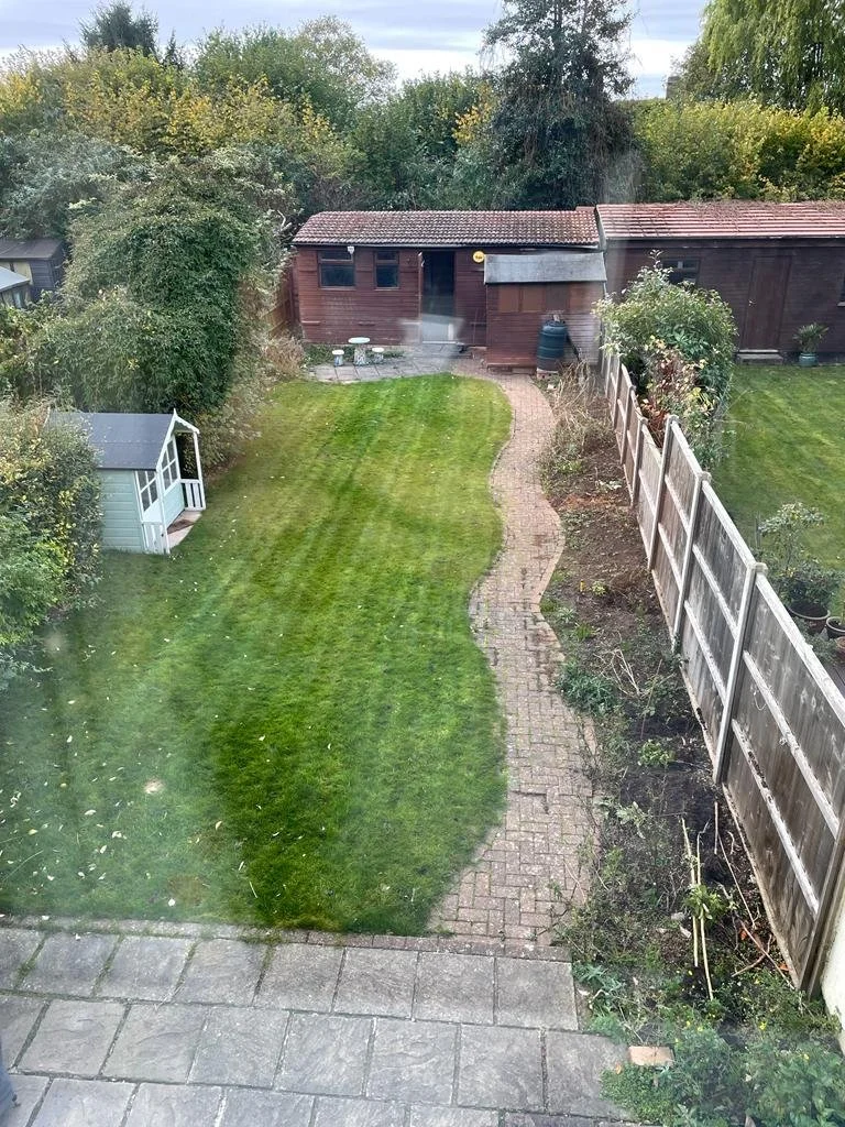 A backyard with a paved patio, a curving brick walkway, a green lawn, and a wooden shed at the back, surrounded by trees and fencing.