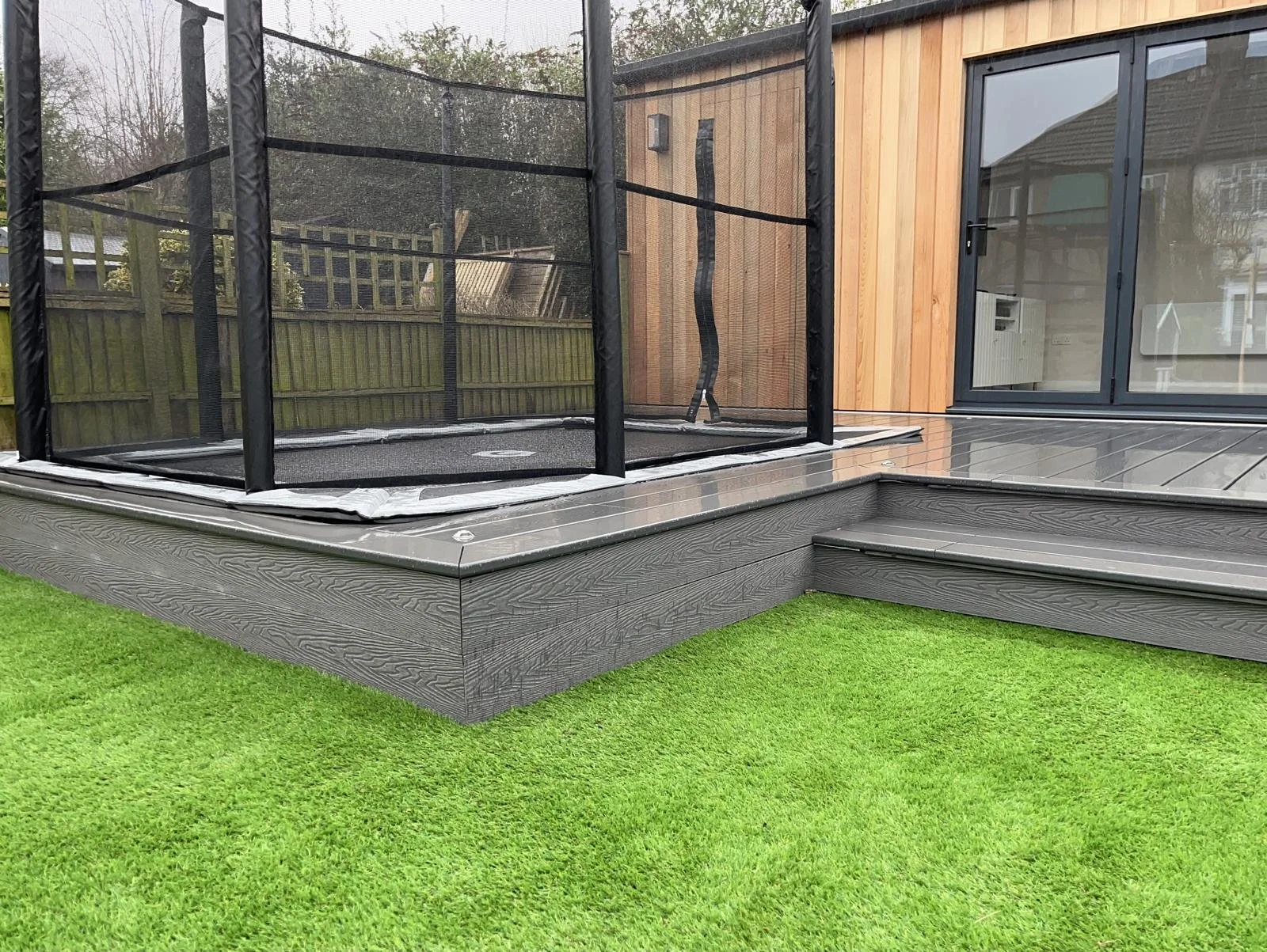 Backyard patio with a trampoline enclosed with black mesh, wooden deck, and sliding glass door leading into a house, with green grass in the foreground.