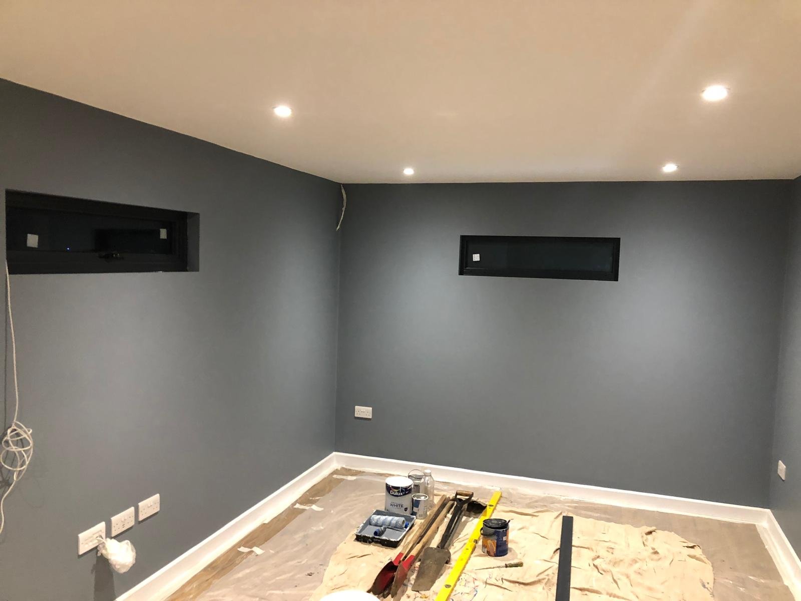 Room under renovation with dark gray painted walls, small horizontal windows, ceiling lights, and tools on the floor covered with protective paper.
