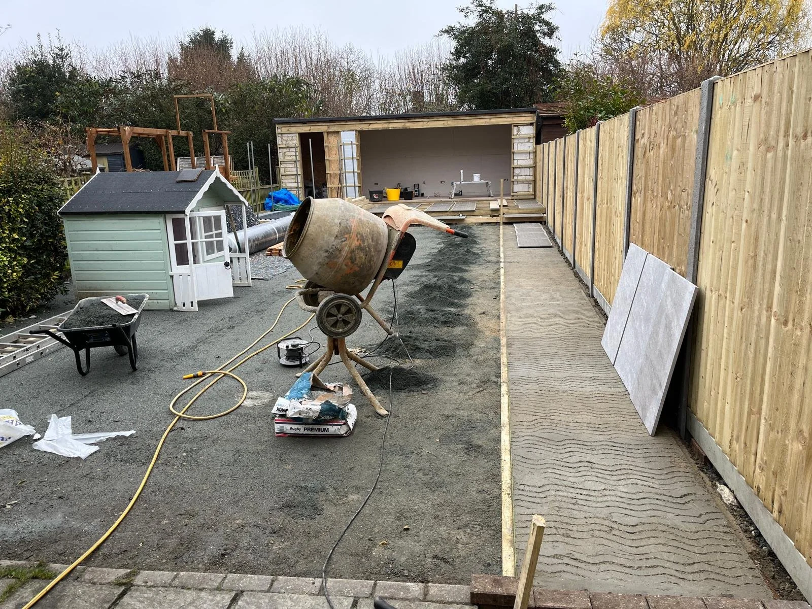 Backyard under construction with a small shed, a concrete mixer, construction tools, and materials. A new wooden fence is being installed along the right side.