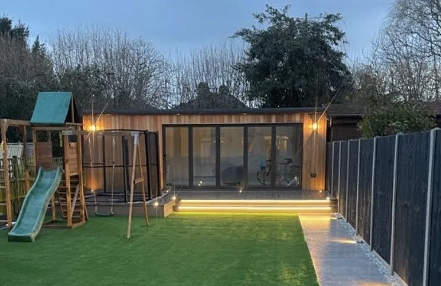 Backyard with a wooden shed, large sliding glass door, outdoor lights, children's play area with slide, and a trampoline, during dusk.