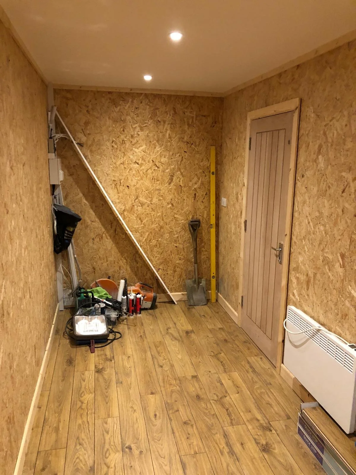 A small room with wood-paneled walls and flooring, containing construction tools and equipment, including a shovel, level, power tools, and a work light, indicating ongoing renovation.