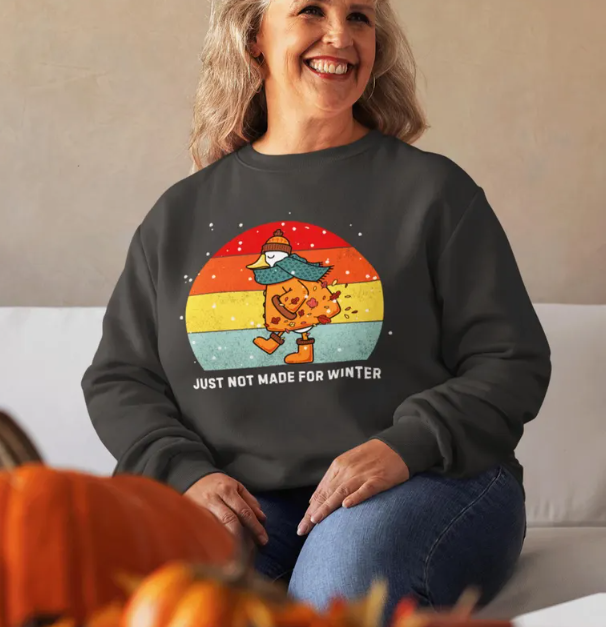 A smiling woman wearing a dark gray sweatshirt with a colorful graphic of a cat wearing a winter hat and scarf, holding a cup, with a rainbow background and the text 'Just Not Made for Winter.'