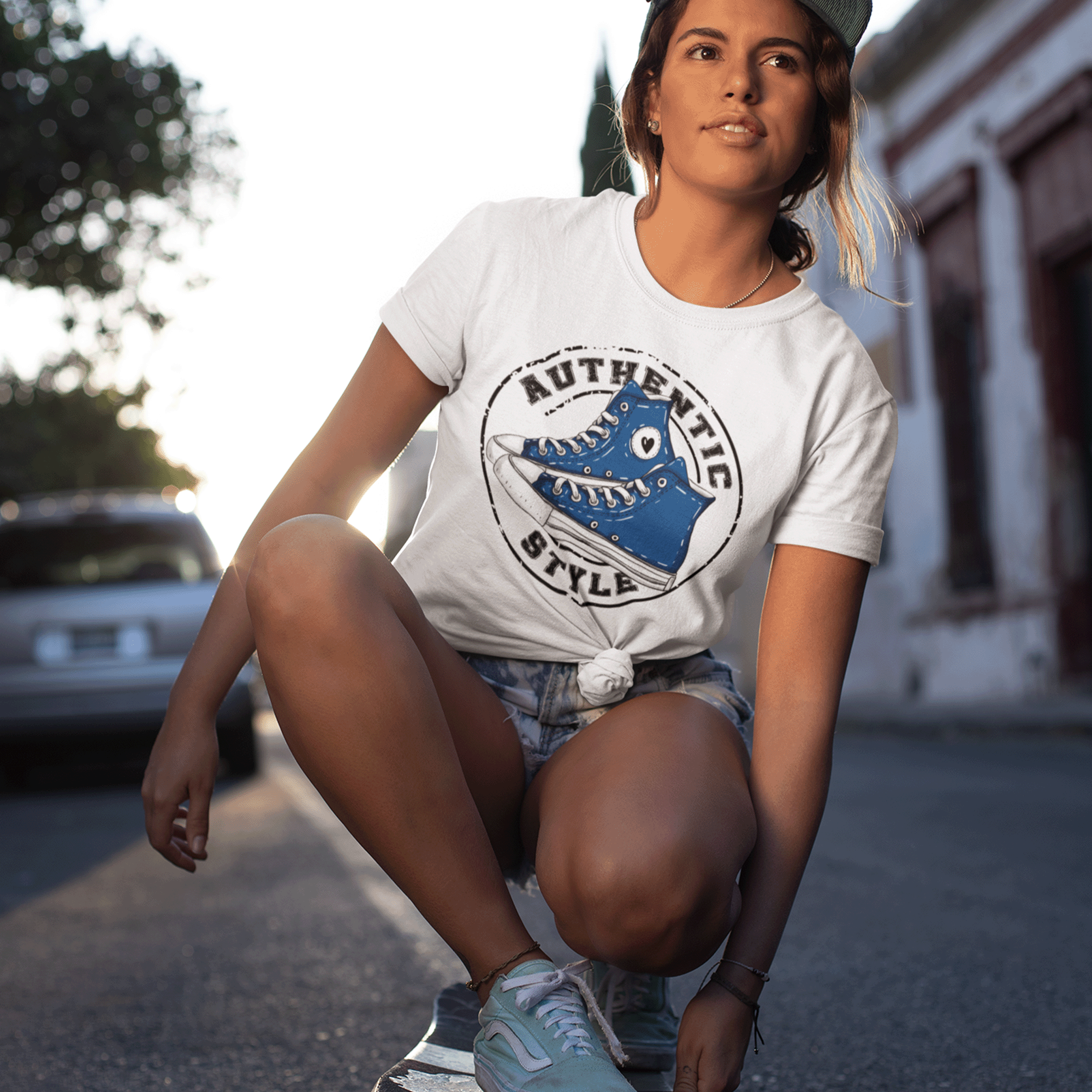 A young woman crouching on a street, wearing sneakers and a white t-shirt with a graphic of a blue roller skate. She has bracelets on her wrist, earrings, and a baseball cap worn backward. The background shows blurred trees and buildings with sunlight creating a backlit effect.