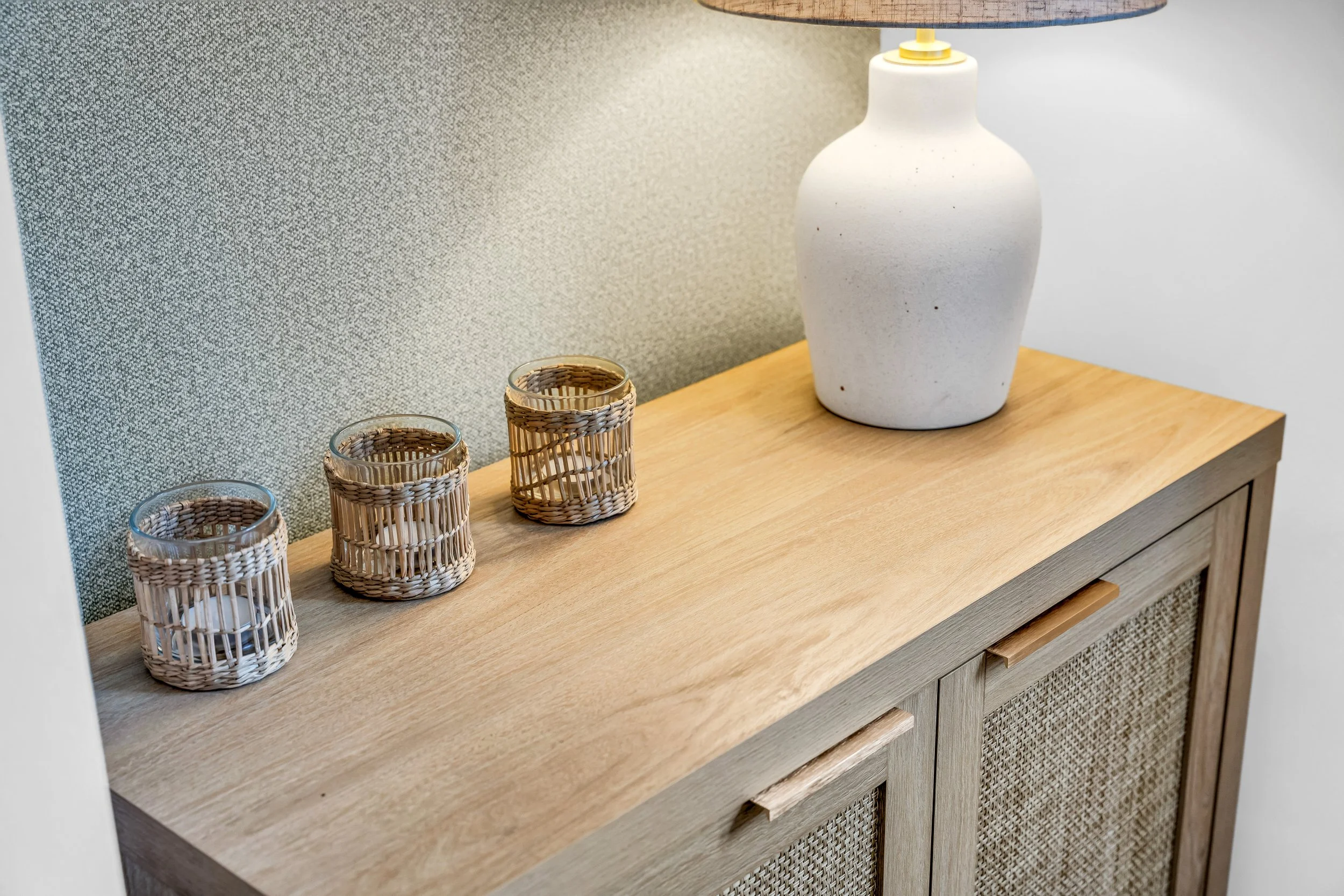 A wooden sideboard with four wicker candle holders and a large white ceramic lamp with a wooden base against a textured beige wall.