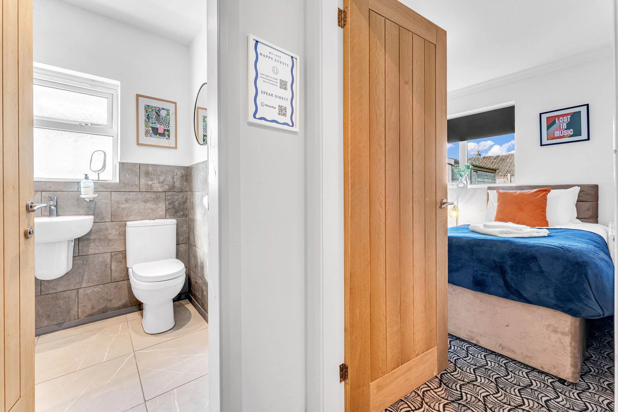 Interior view of a bedroom with a bed featuring a blue blanket and orange pillow, and a bathroom with a toilet and sink, separated by a wooden door, with decorative art on the walls.