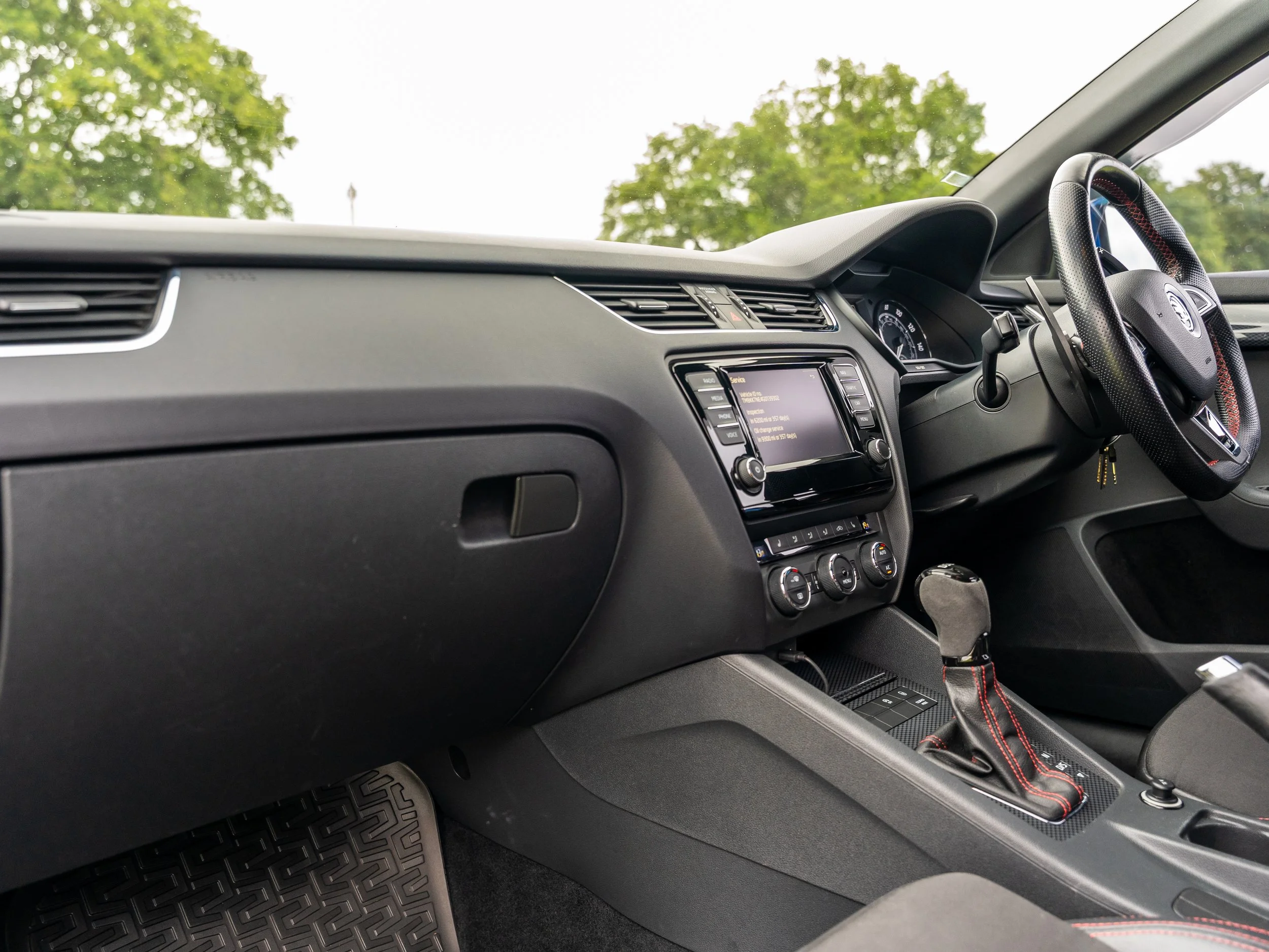 Interior of a car showing dashboard, steering wheel, gear shift, and infotainment system, with trees visible outside the window.