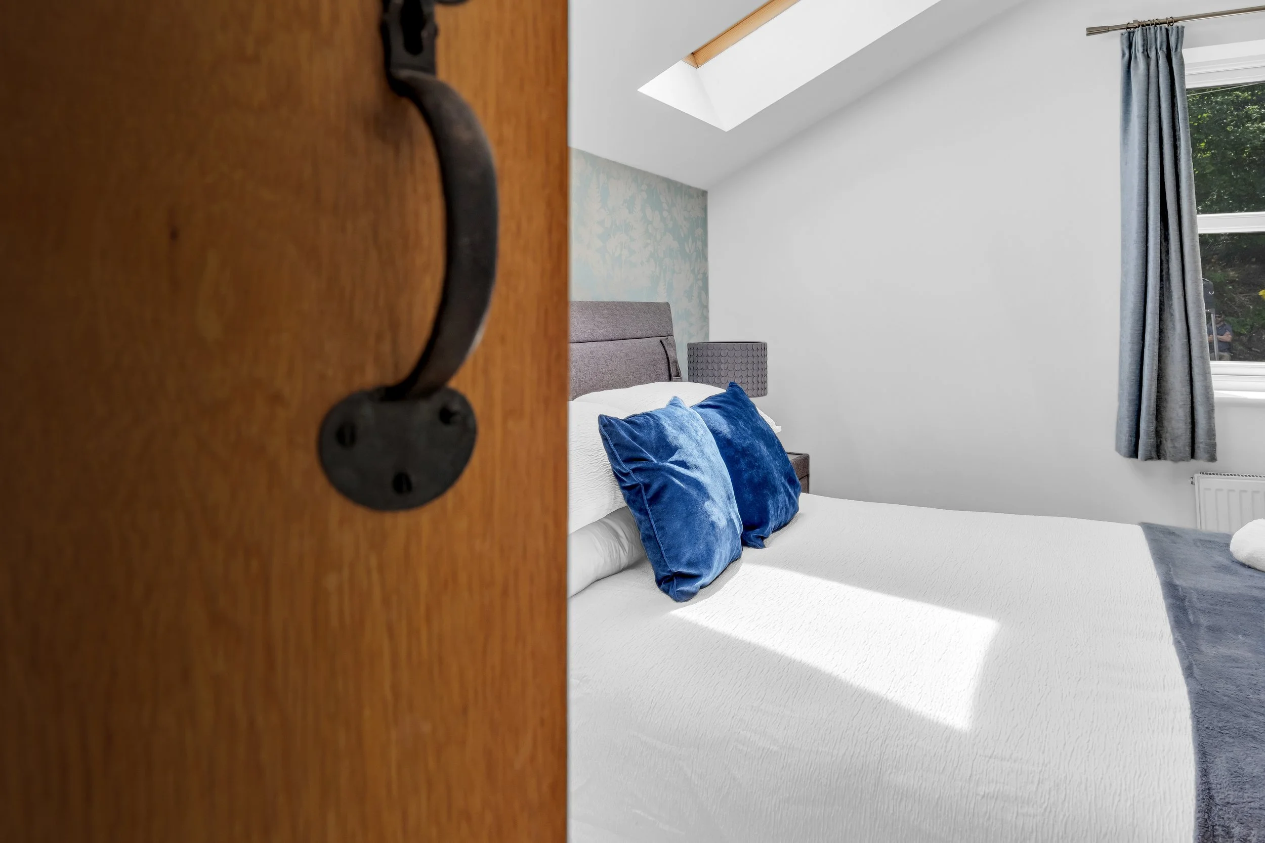 View of a bedroom with white walls, a bed with blue pillows, a window with blue curtains, and a skylight in the ceiling, seen through an open wooden door.