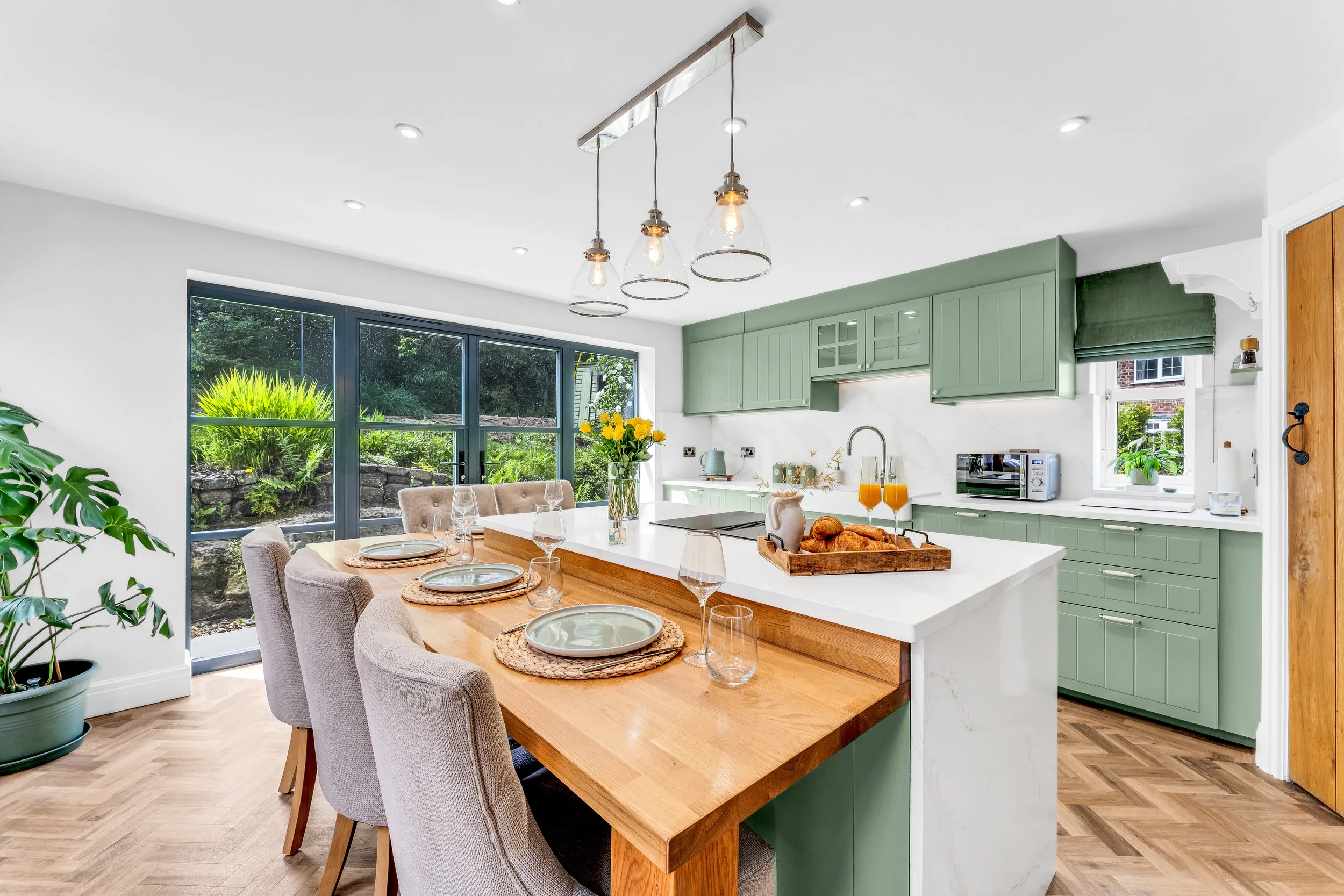 A bright and airy kitchen and dining area with a large window overlooking a garden. The space features green cabinetry, a white marble backsplash, a wooden dining table with beige upholstered chairs, and modern pendant lighting.